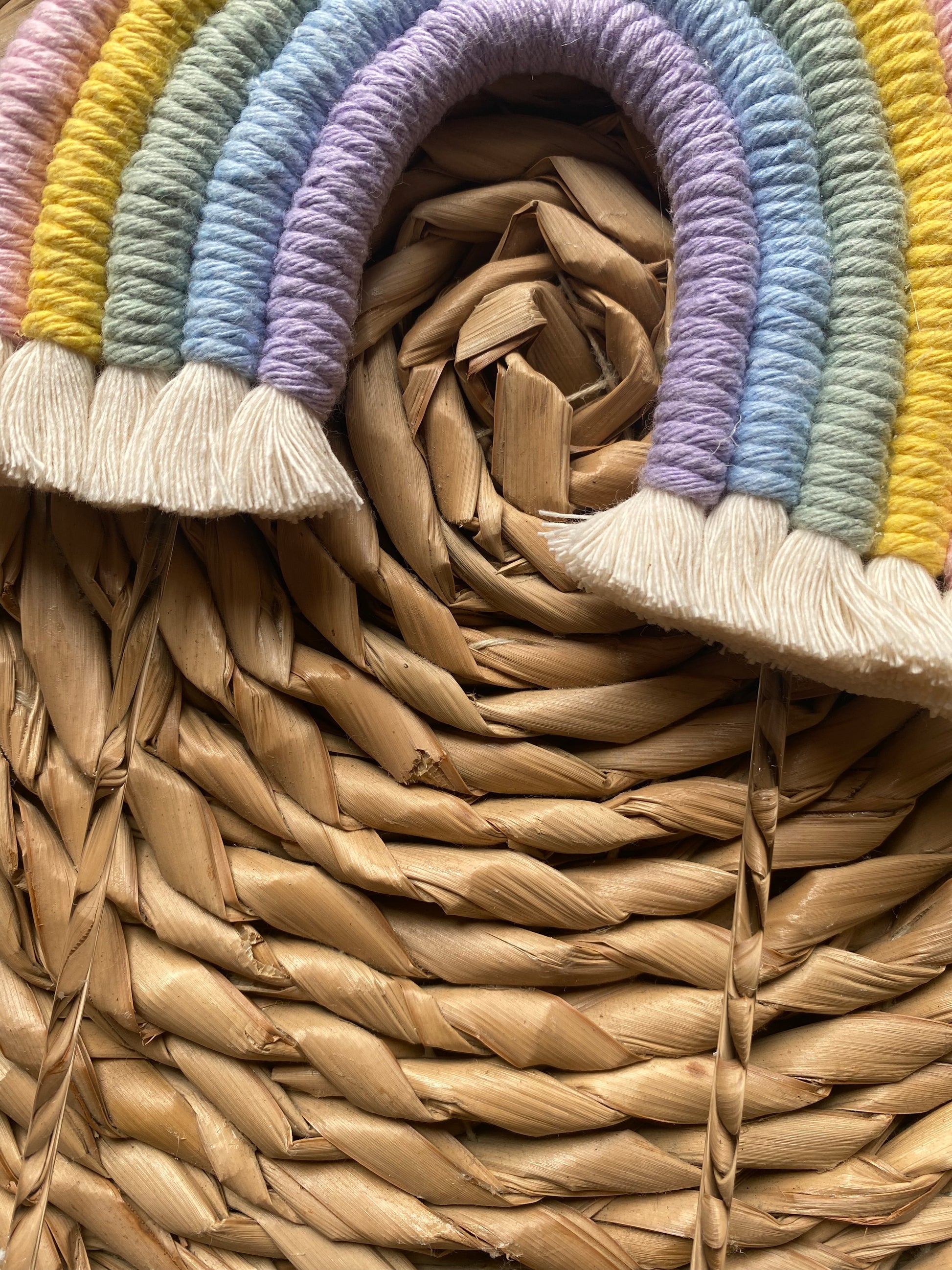 Close up of a pastel rainbow macramé cake topper with long clear acrylic sticks, laid on a round wicker mat.