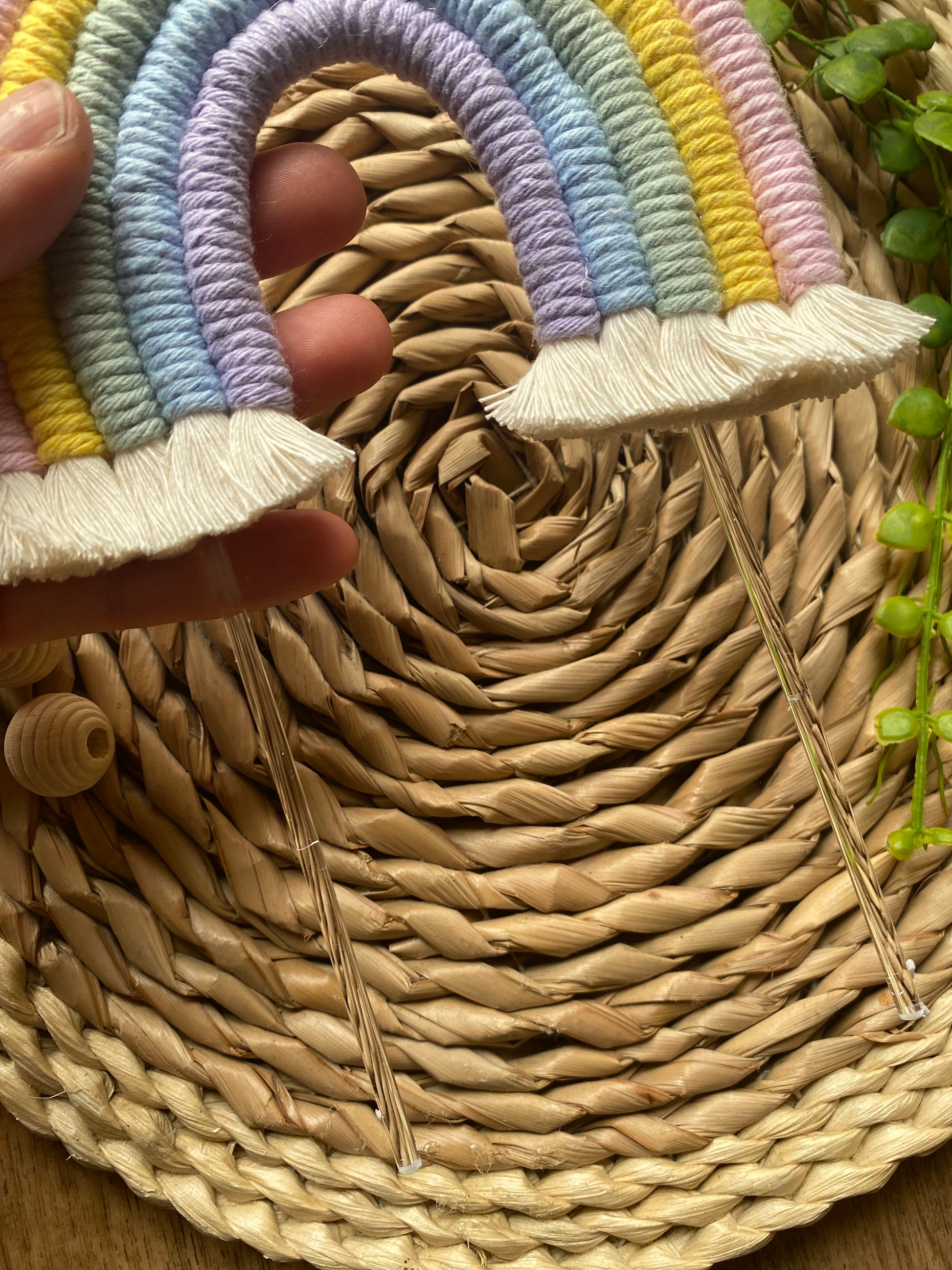 Pastel rainbow macramé cake topper with long clear acrylic sticks, held in a persons hand over a round wicker mat.