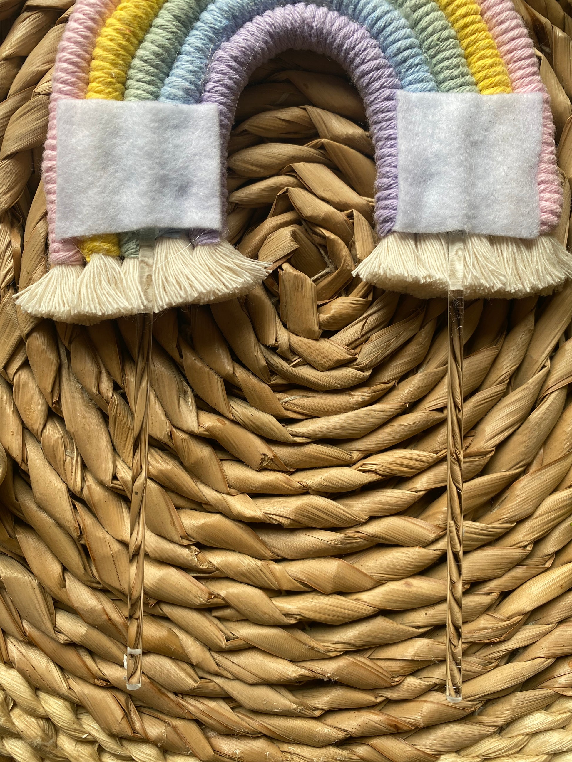 Reverse side of a pastel rainbow macramé cake topper with long clear acrylic sticks, laid on a round wicker mat. The rainbow has white felt patches where the acrylic sticks are attached.