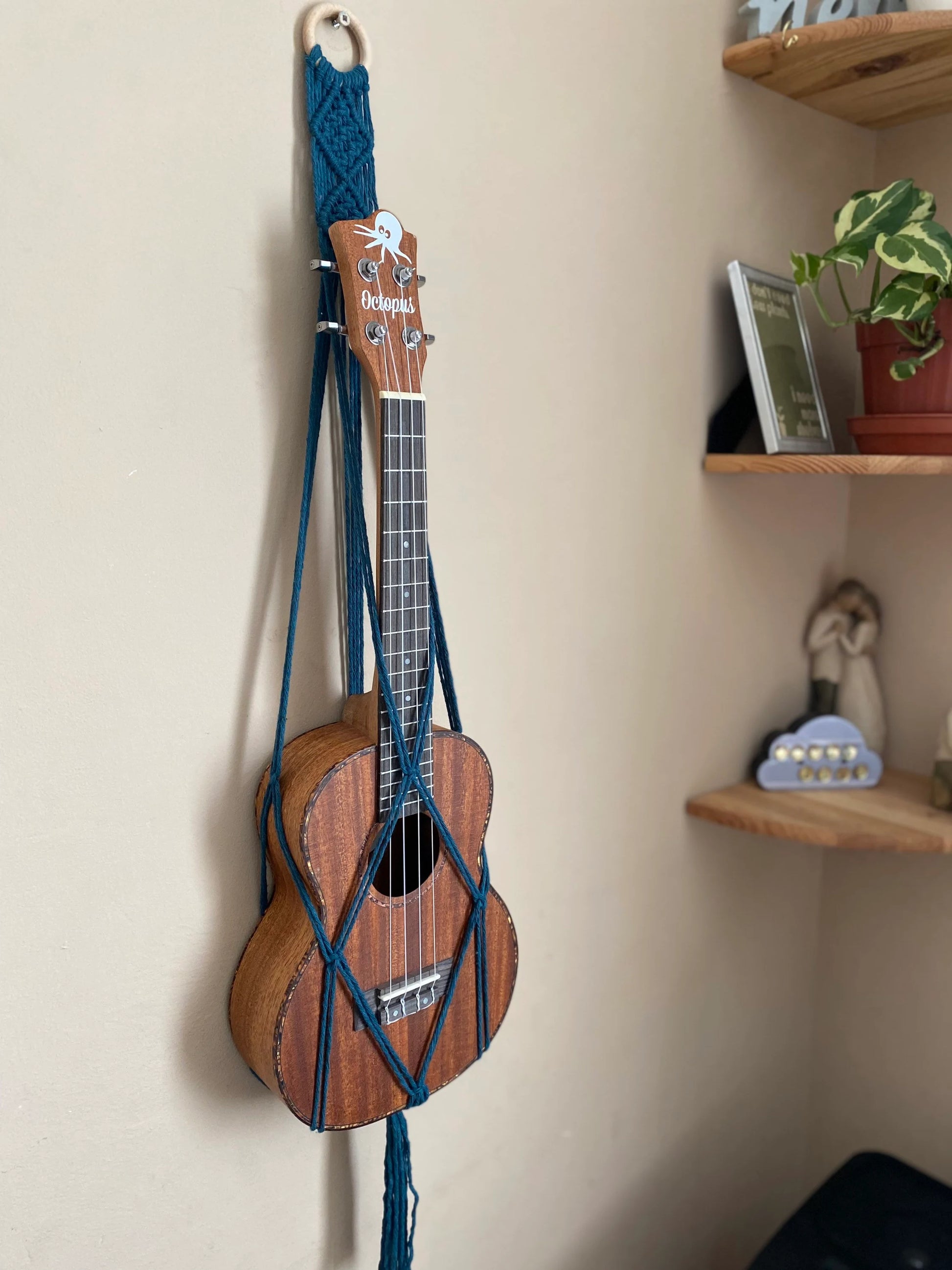 A dark brown ukulele sits inside of a handmade macrame wall hanging ukulele hanger, which is made with petrol blue recycled cotton, and a repurposed wooden hanging ring.