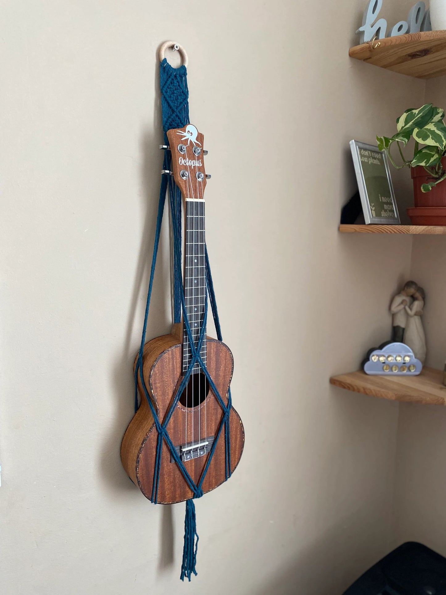 A dark brown ukulele sits inside of a handmade macrame wall hanging ukulele hanger, which is made with petrol blue recycled cotton, and a repurposed wooden hanging ring. A set of corner shelves is to the right of the image.