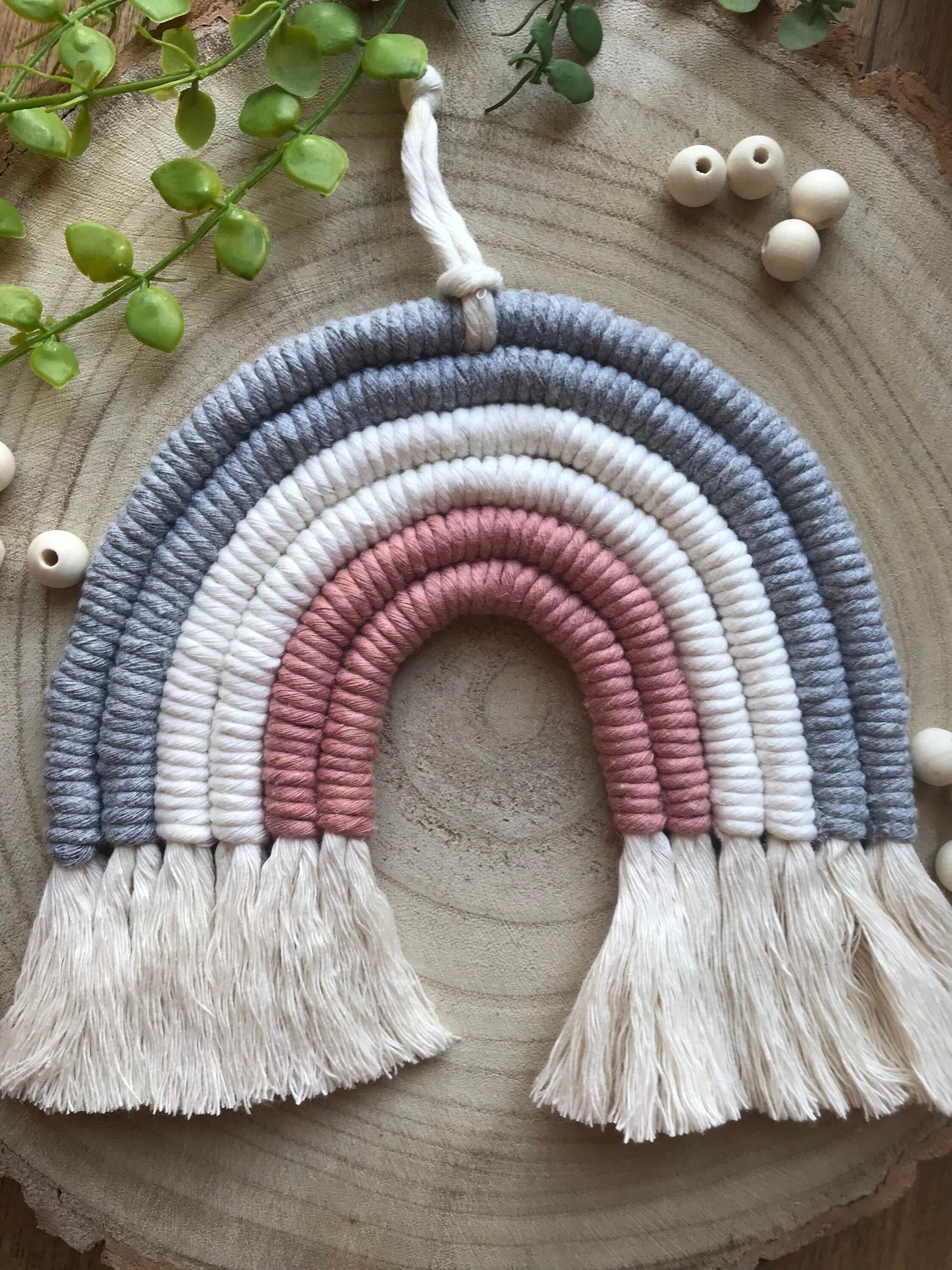 A macrame rainbow made with 6 arches of grey, natural and pink yarn with combed out tassels at the bottom, displayed on a wooden board with green plant vines and wooden beads.