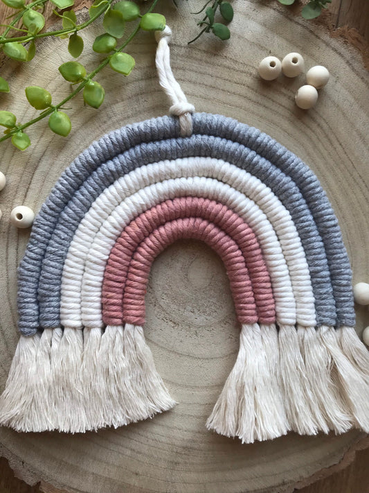 A macrame rainbow made with 6 arches of grey, natural and pink yarn with combed out tassels at the bottom, displayed on a wooden board with green plant vines and wooden beads.