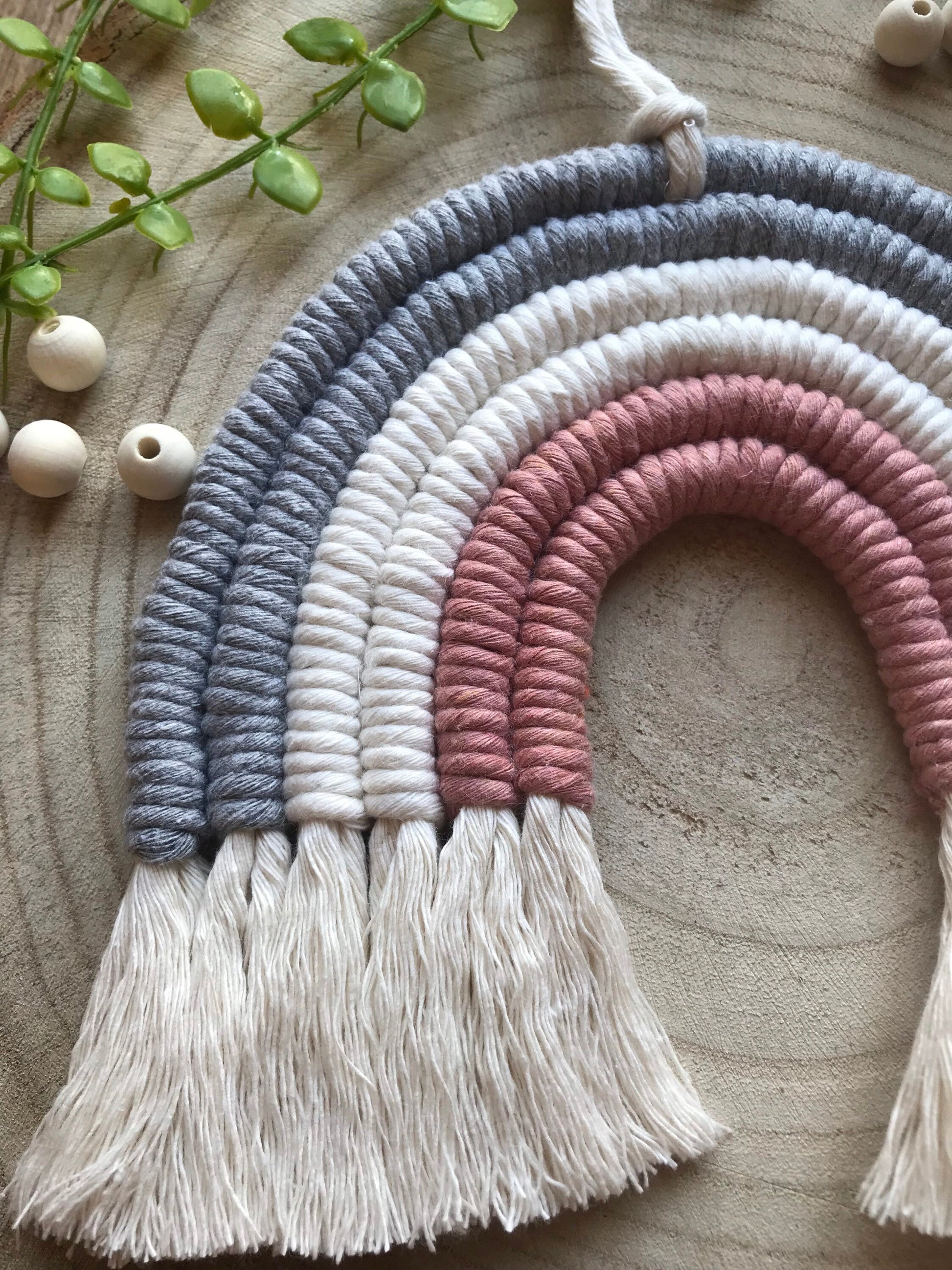 Close up of a macrame rainbow made with 6 arches of grey, natural and pink yarn with combed out tassels at the bottom, displayed on a wooden board with green plant vines and wooden beads.