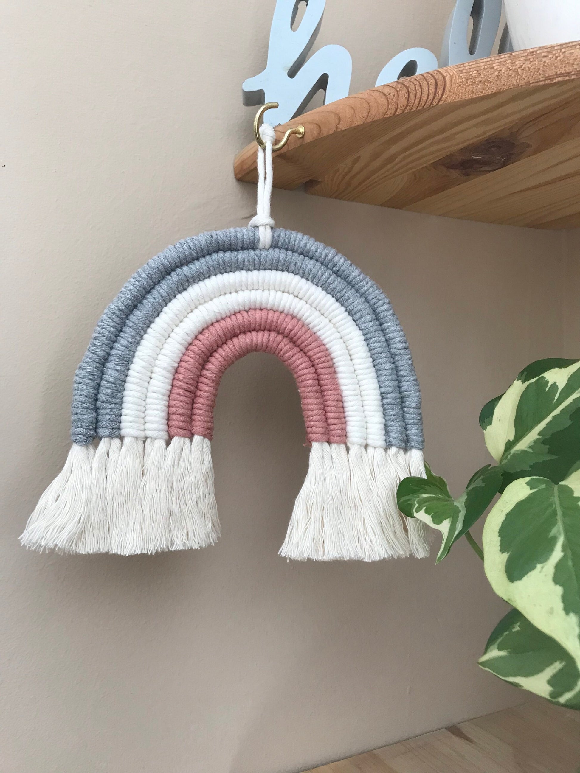 A macrame rainbow made with 6 arches of grey, natural and pink yarn with combed out tassels at the bottom, hanging from a wooden corner shelf next to a variegated houseplant.
