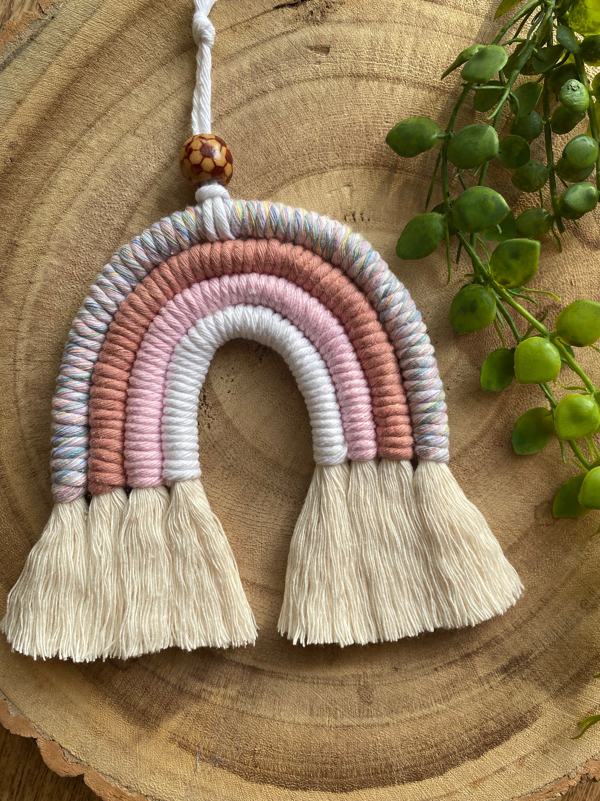 A handmade macrame rainbow with 4 arches, made with pink and white tones of cotton yarn, finished with a patterned wooden bead at the top, and natural combed out fringe at the bottom.