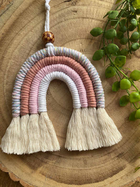 A handmade macrame rainbow with 4 arches, made with pink and white tones of cotton yarn, finished with a patterned wooden bead at the top, and natural combed out fringe at the bottom.