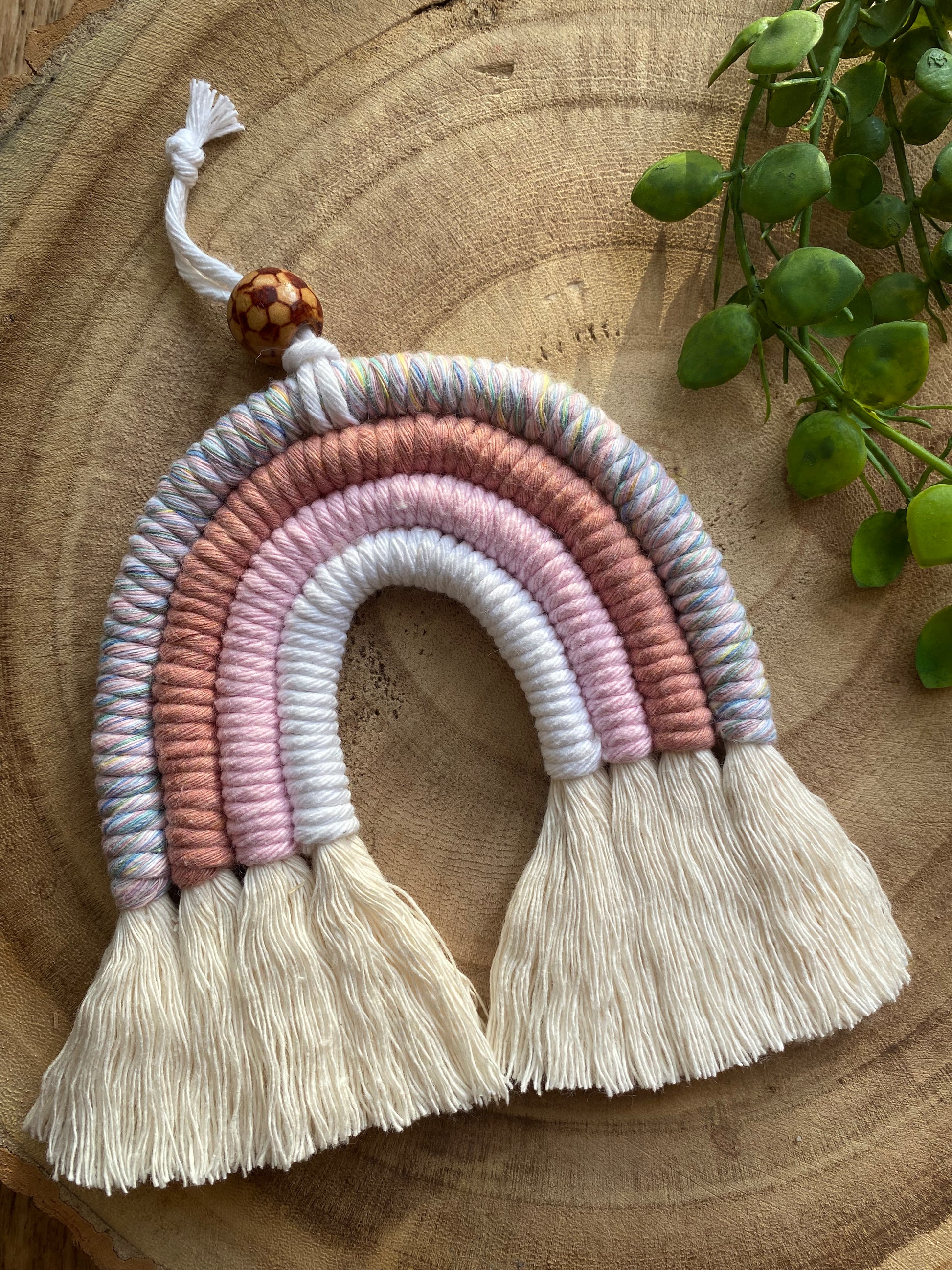 A top down view of a handmade macrame rainbow with 4 arches, made with pink and white tones of cotton yarn, finished with a patterned wooden bead at the top, and natural combed out fringe at the bottom.