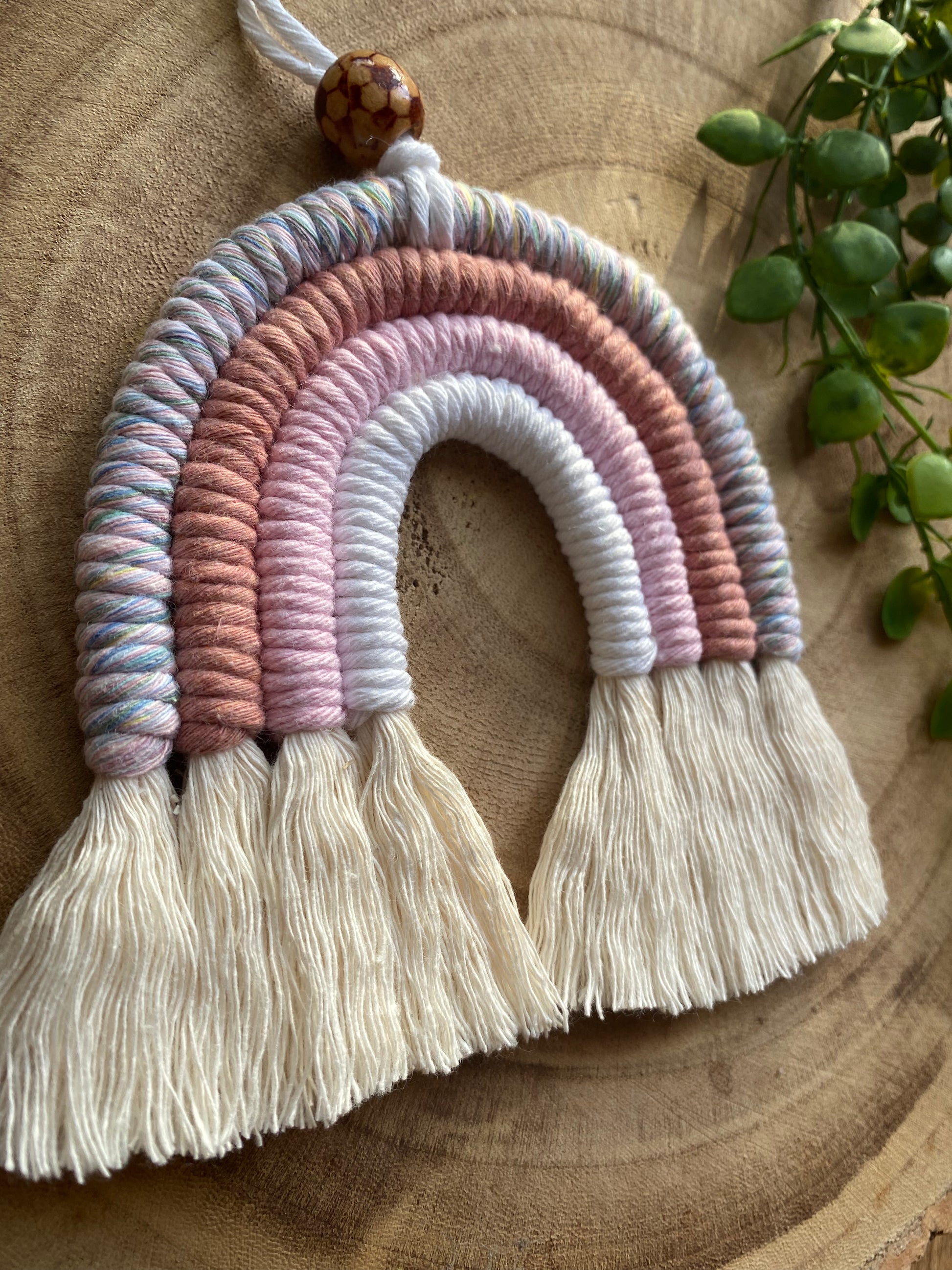 A close up of a handmade macrame rainbow with 4 arches, made with pink and white tones of cotton yarn, finished with a patterned wooden bead at the top, and natural combed out fringe at the bottom.