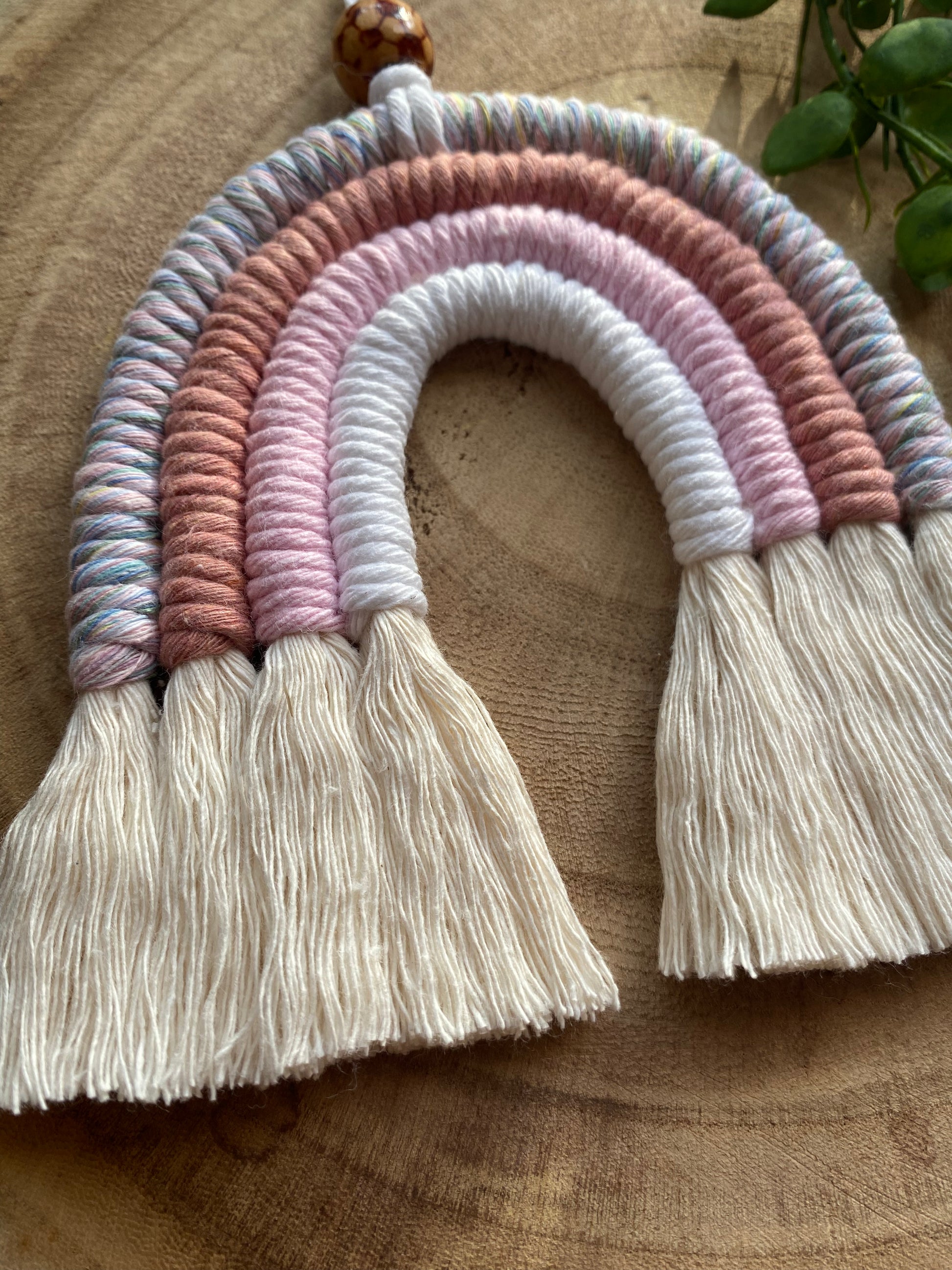 Close up of a handmade macrame rainbow with 4 arches, made with pink and white tones of cotton yarn, finished with a patterned wooden bead at the top, and natural combed out fringe at the bottom.
