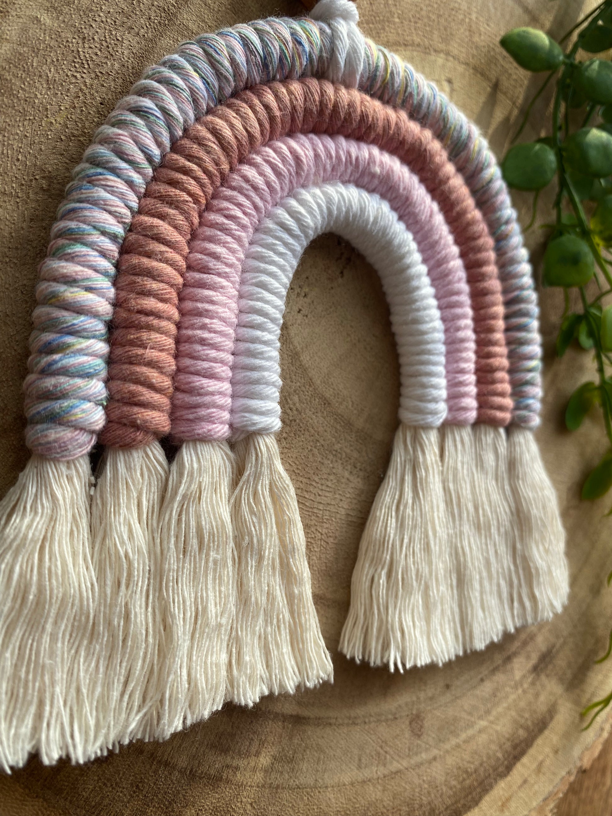 A handmade macrame rainbow with 4 arches, made with pink and white tones of cotton yarn, and natural combed out fringe at the bottom, lays on a wooden surface.