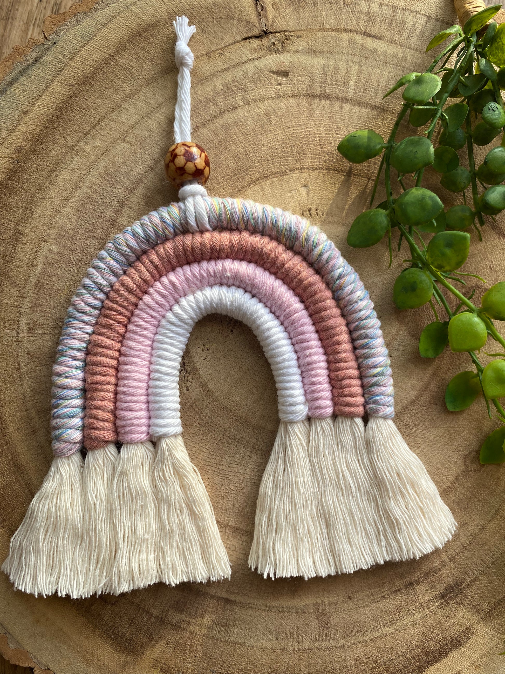 A handmade macrame rainbow with 4 arches, made with pink and white tones of cotton yarn, finished with a patterned wooden bead at the top, and natural combed out fringe at the bottom.