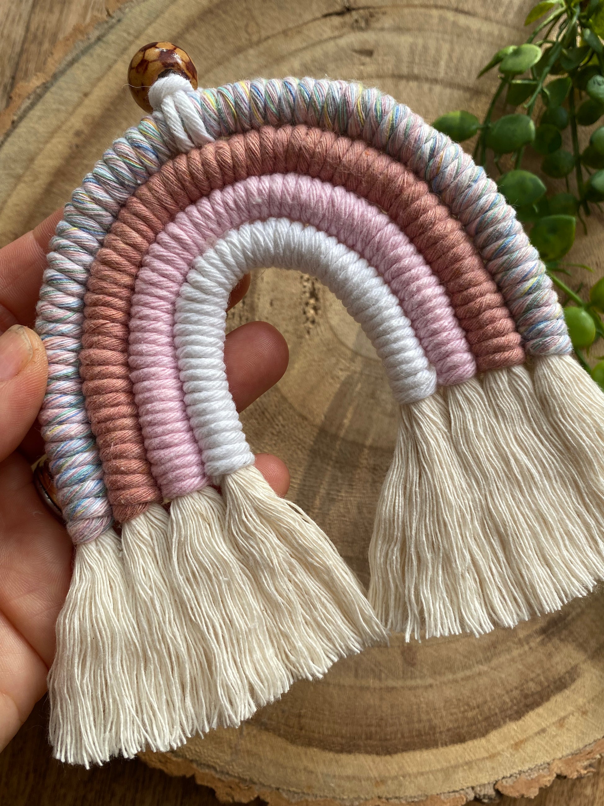 A hand holding a handmade macrame rainbow with 4 arches, made with pink and white tones of cotton yarn, finished with a patterned wooden bead at the top, and natural combed out fringe at the bottom.
