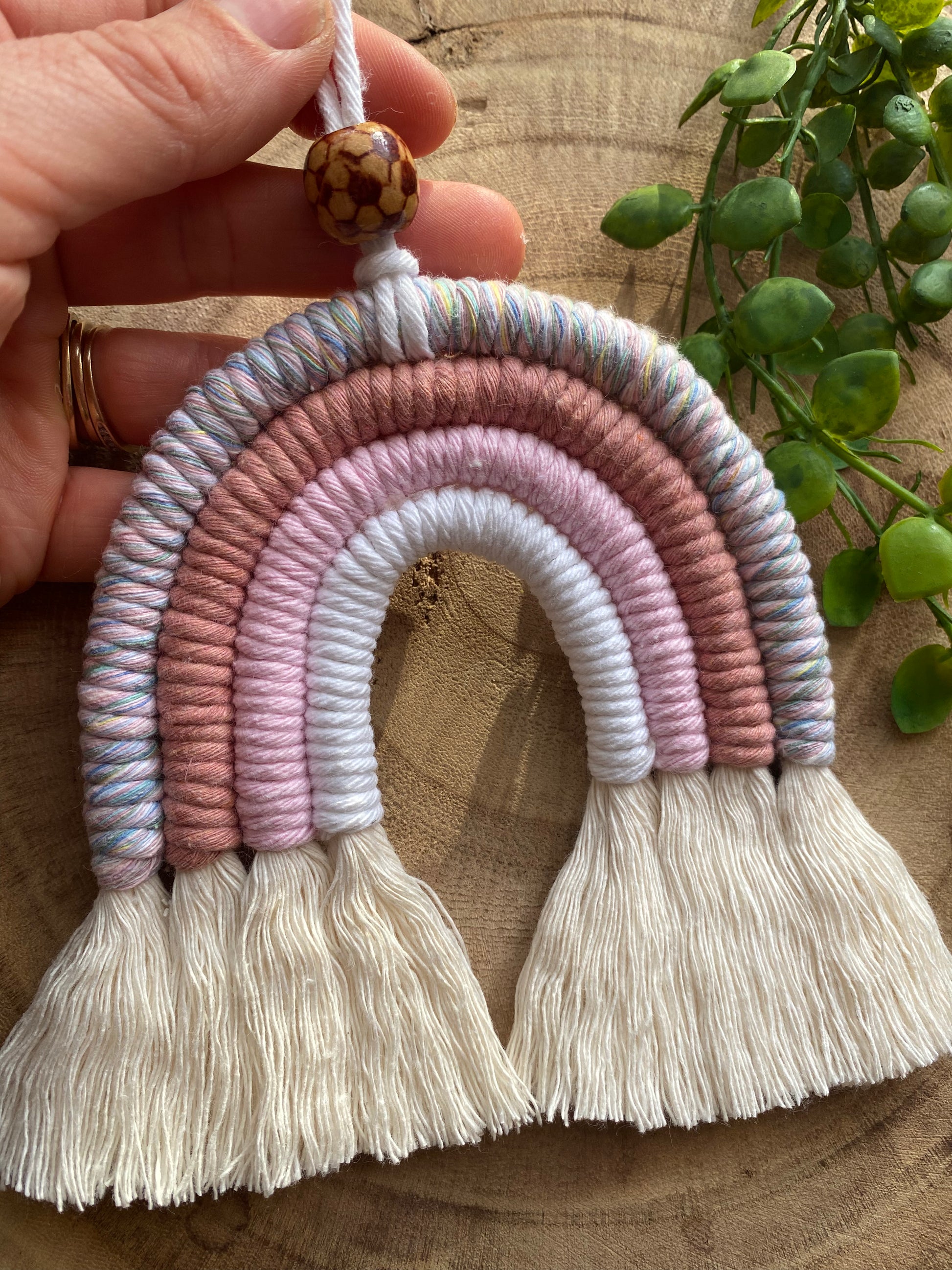 A hand holding the hanging cord at the top of a handmade macrame rainbow with 4 arches, made with pink and white tones of cotton yarn, finished with a patterned wooden bead at the top, and natural combed out fringe at the bottom.