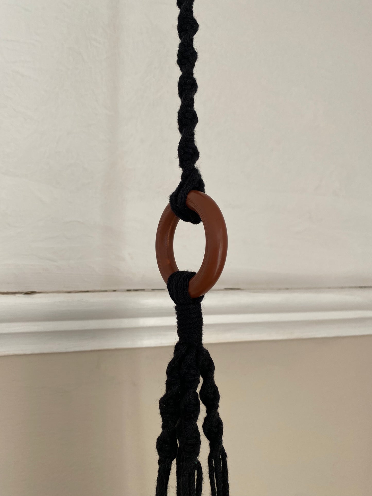 A macrame plant hanger extension made with black cotton yarn, is attached to the wooden ring of a macrame plant hanger.