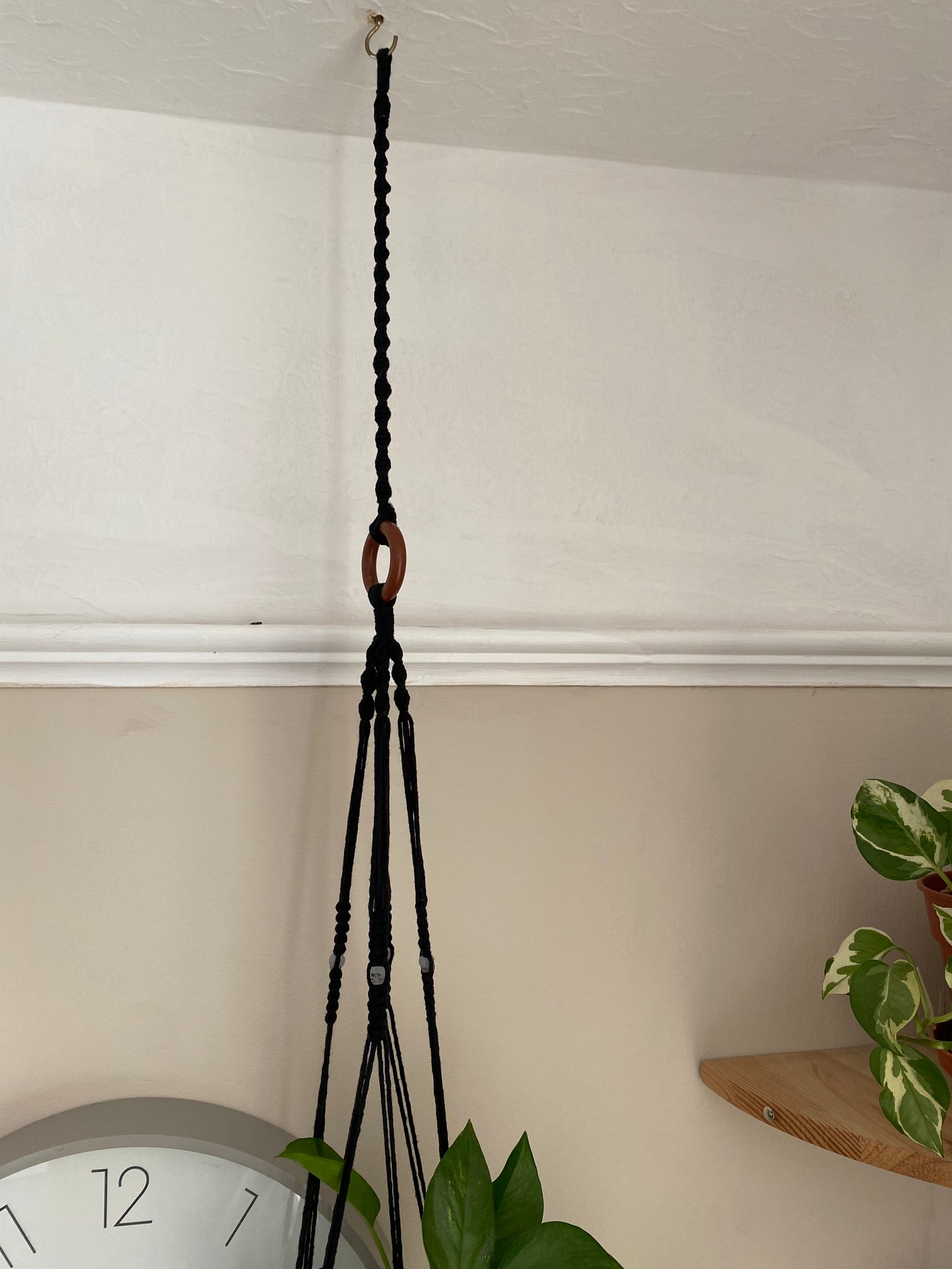A macrame plant hanger extension made with black cotton yarn, is attached to the wooden ring of a macrame plant hanger, and suspended from a white ceiling on a metal hook. You can see the edges of two houseplants in the bottom and right of the image, and the plant hanger has small skull beads on it.