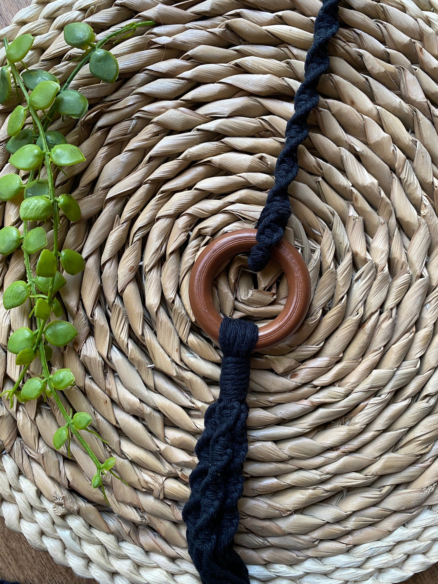 A macrame plant hanger extension made with black cotton yarn, is attached to the wooden ring of a macrame plant hanger, and laid across a wicker mat.