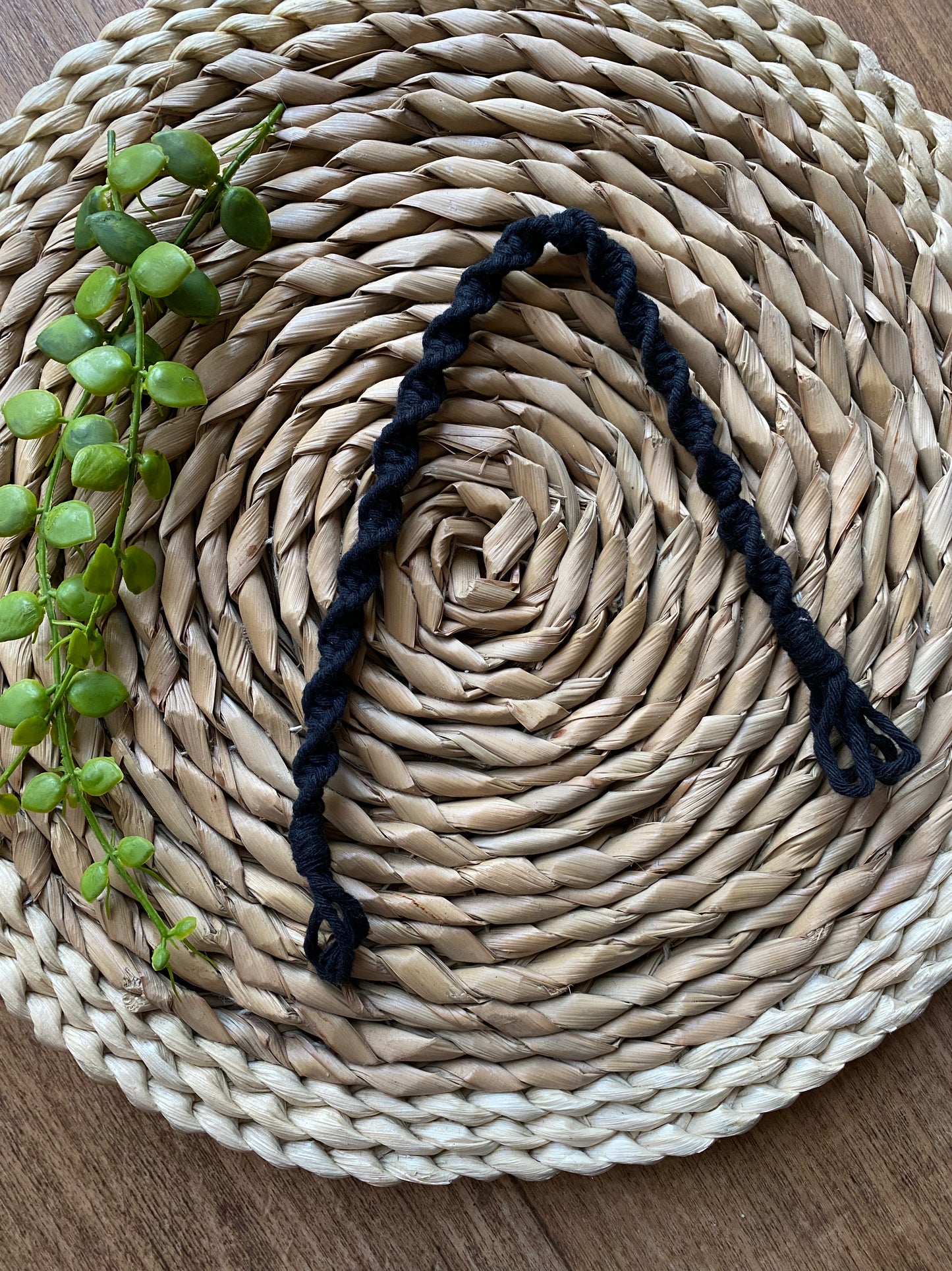 A macrame plant hanger extension made with black cotton yarn, is folded on a round wicker mat.