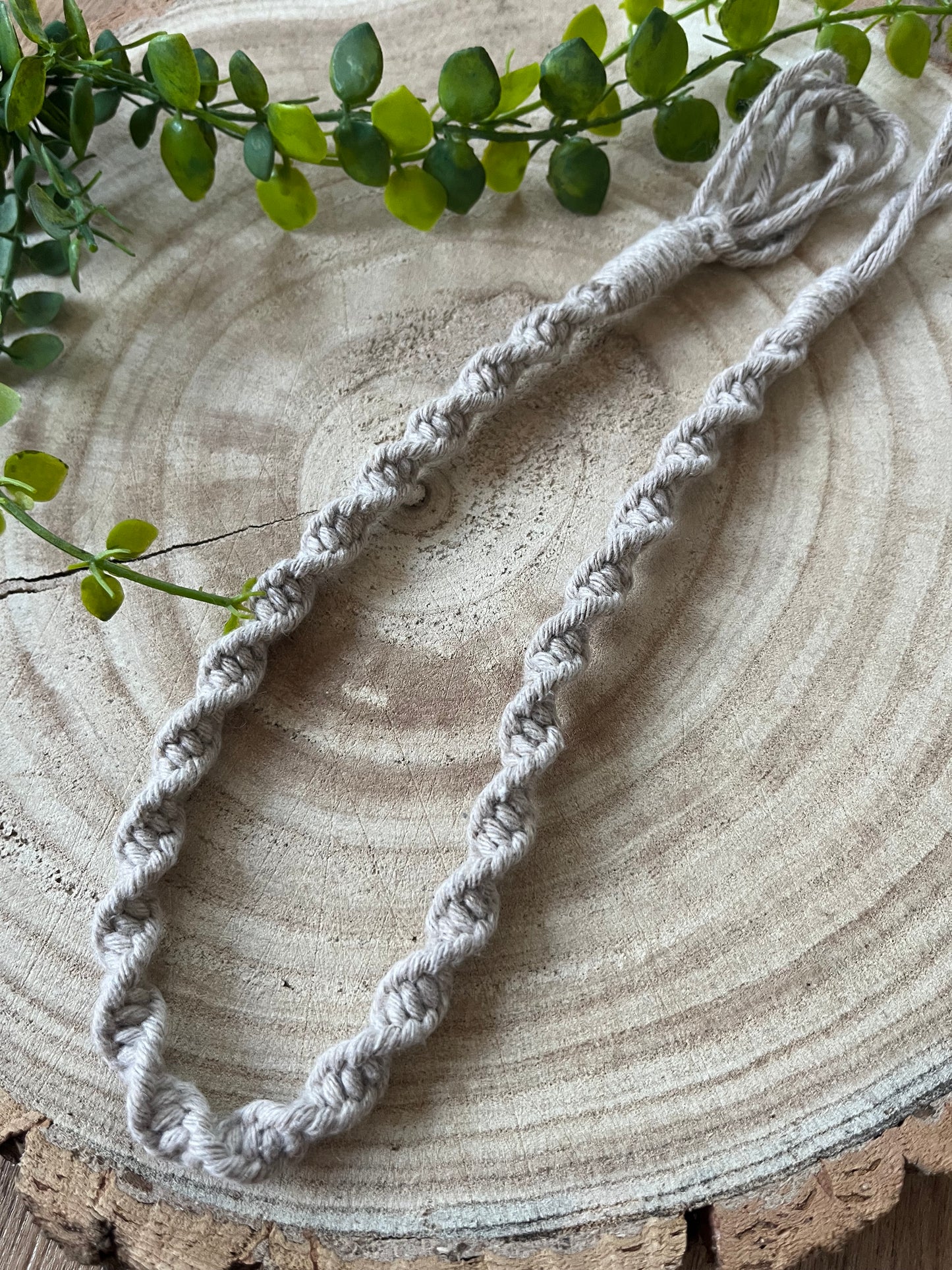 A handmade macramé plant hanger extension from Macra-Made-With-Love sits on a textured wooden surface, adorned with green leaves. Crafted from recycled cotton, the plant hanger extension is intricately knotted in a spiral pattern and made of taupe-coloured cord.