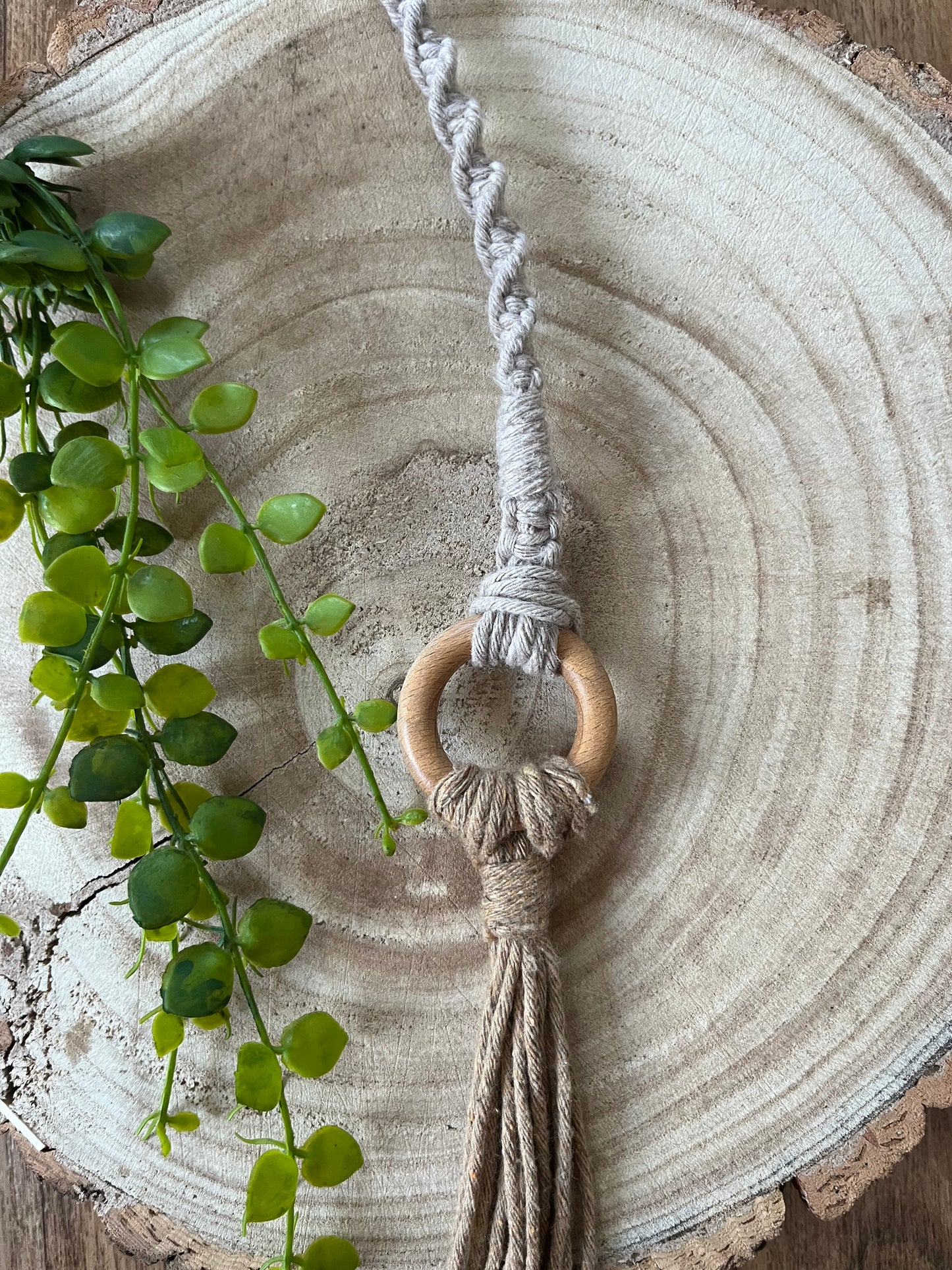 A plant hanger extension from Macra-Made-With-Love lies on a wooden slab attached to the wooden ring of a macrame plant hanger.