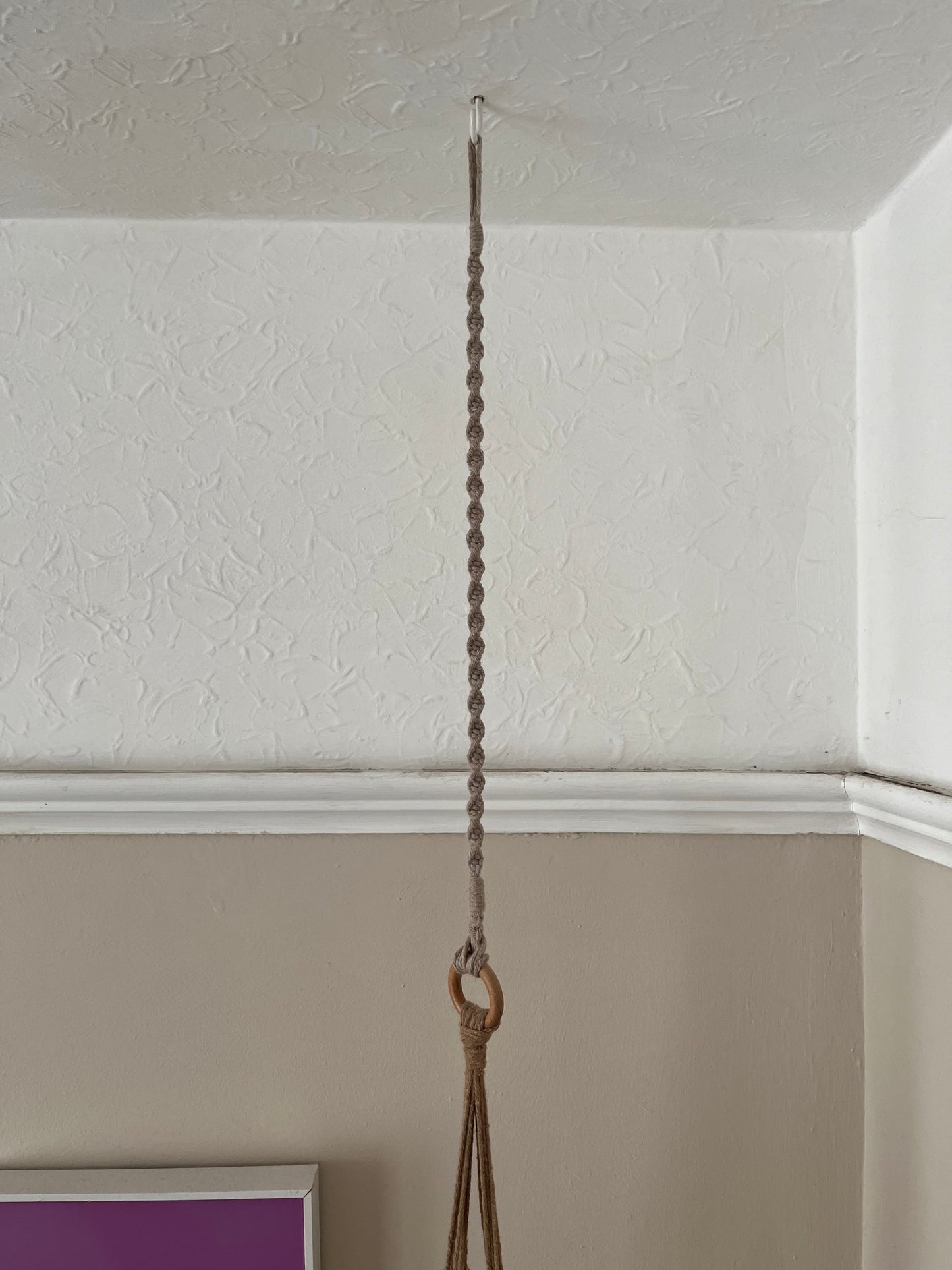 A macrame indoor plant hanger extension, made of taupe, recycled cotton rope, is suspended from a white ceiling hook. Below the hook, the rope is attached to the wooden ring of a macrame plant hanger.