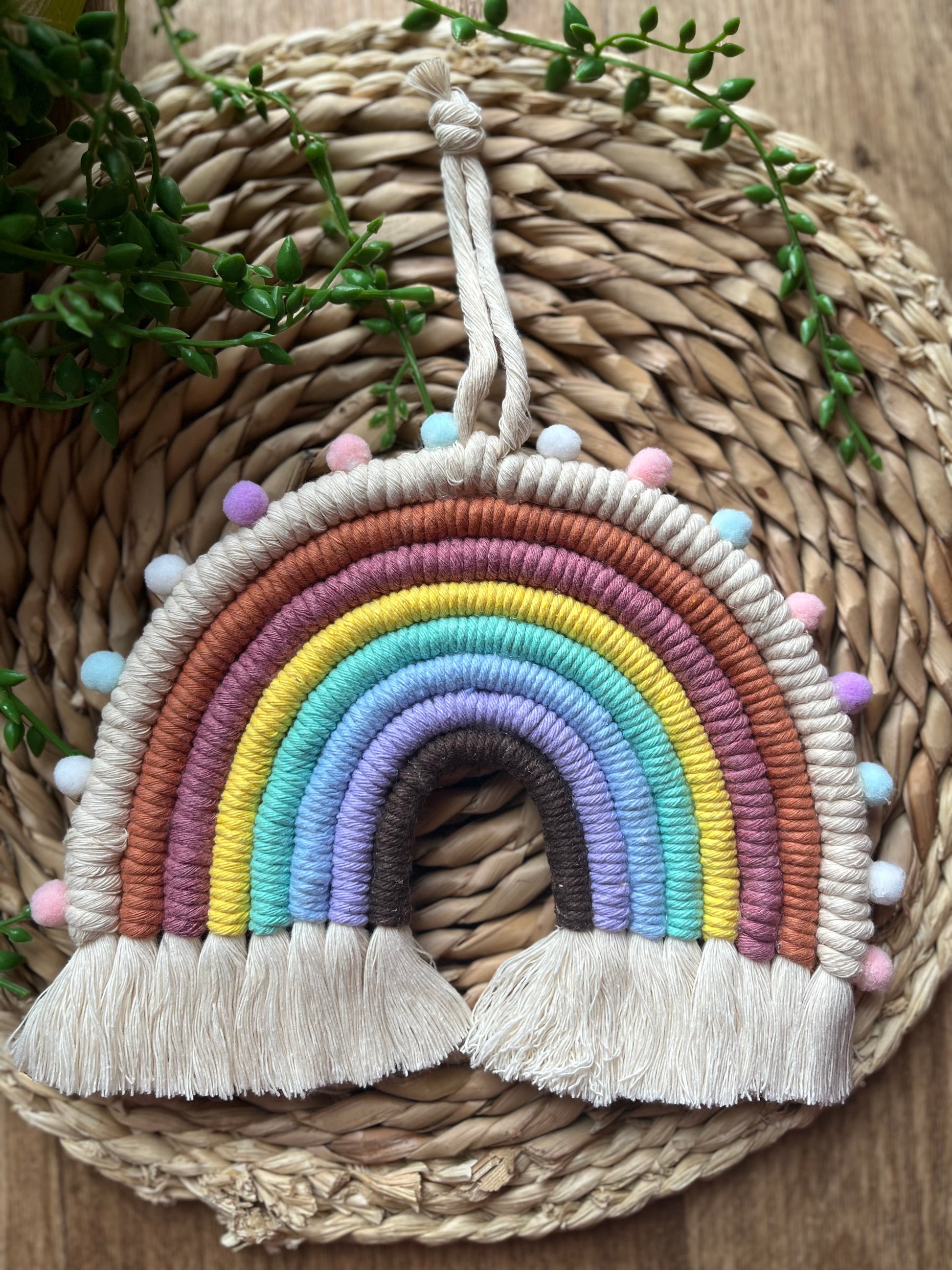 Handmade macrame rainbow wall hanging, made using pastel shades of recycled cotton and finished with small pom poms. laid on a wicker surface with long green plant vines.