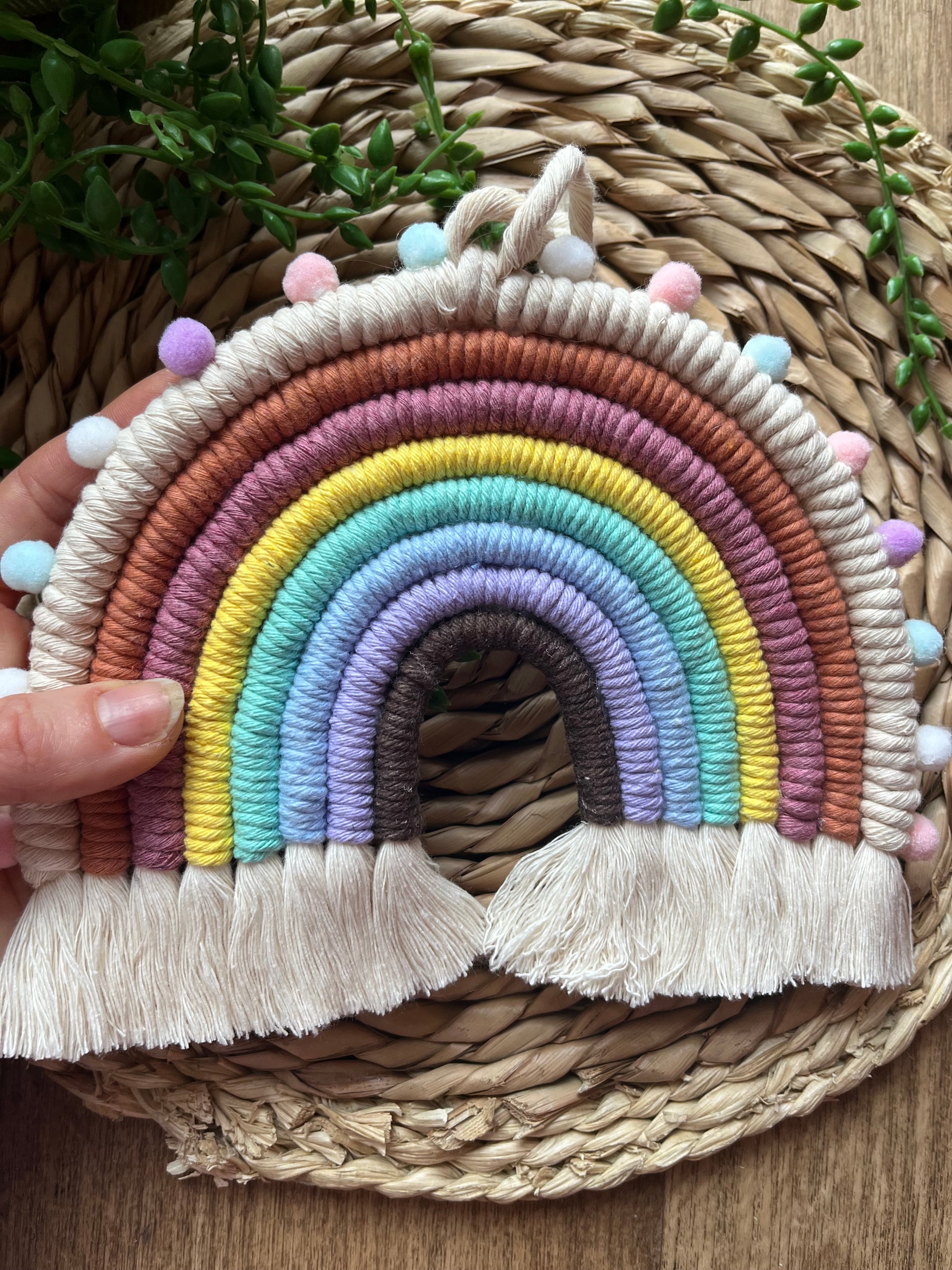 A hand holding a handmade macramé rainbow wall hanging, made with pastel shades of recycled cotton and finished with pom poms.