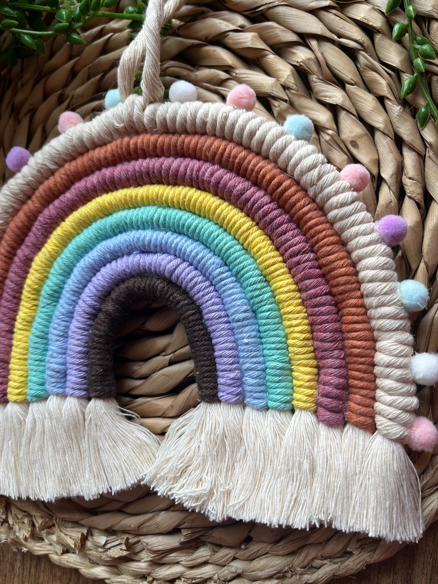 Handmade macrame rainbow wall hanging, made with pastel shades of recycled cotton and finished round the edges with small pom poms.