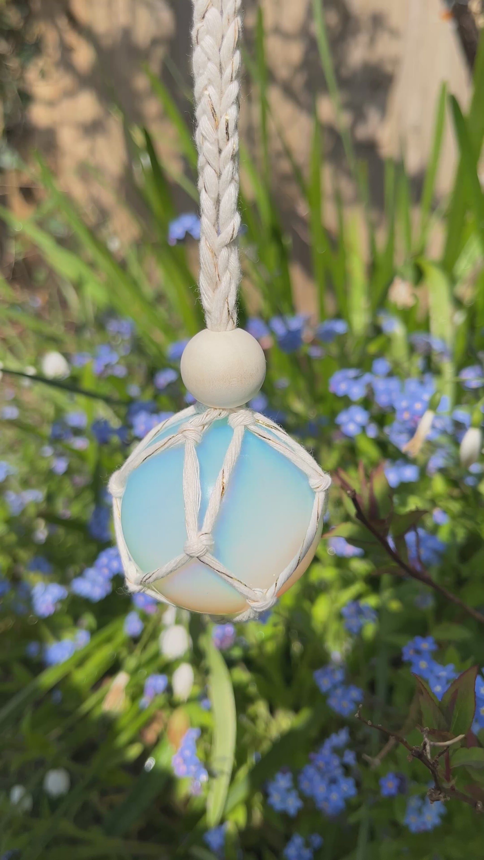 Video showing a handmade macrame opaline sphere hanging sun catcher, made with eco friendly natural recycled cotton yarn. The sphere is shown hanging outside in front of some small blue flowers, there is a plain wooden bead above the crystal.