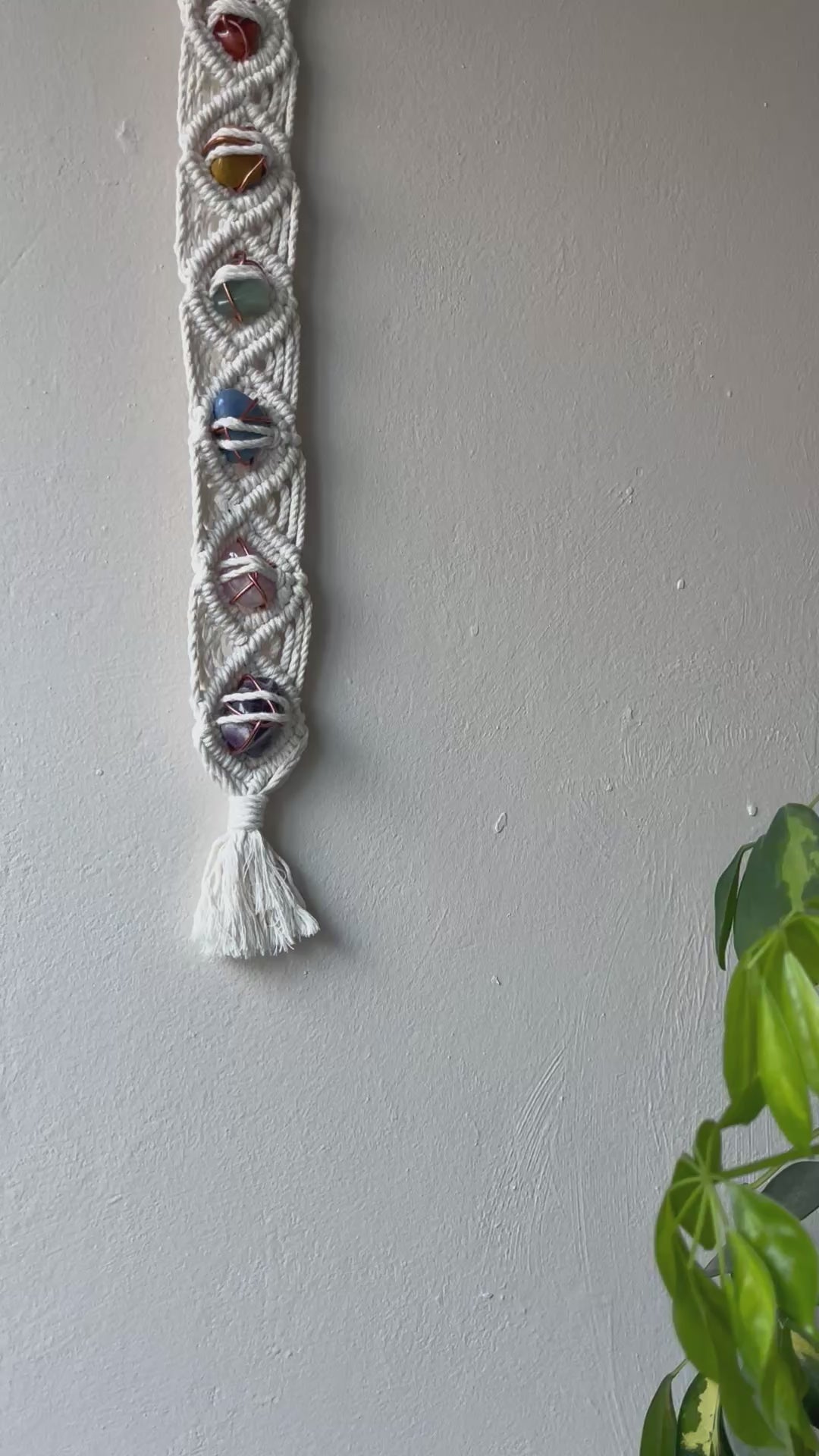 A video panning a long macrame wall hanging, featuring 7 gemstones to represent the chakras, held together with copper wire, and wrapped in natural recycled cotton yarn using a diamond knot pattern. finished with a repurposed wooden hanging ring.