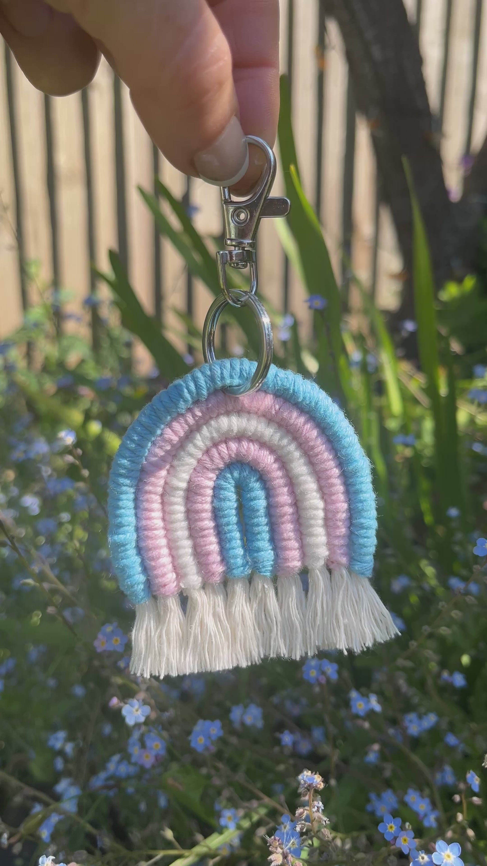 Video showing a handmade macrame rainbow keyring, made with blue, pink and white yarns to represent the colours of the trans pride flag. The keyring has a silver split ring and lobster clasp at the top.