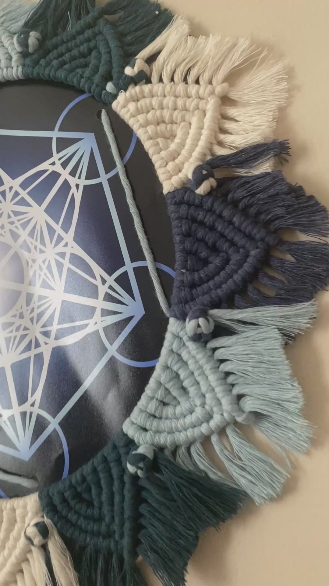 A video showing up close details of a Metatrons Cube Print framed by a Macrame Mandala, knotted with blue and white yarns.