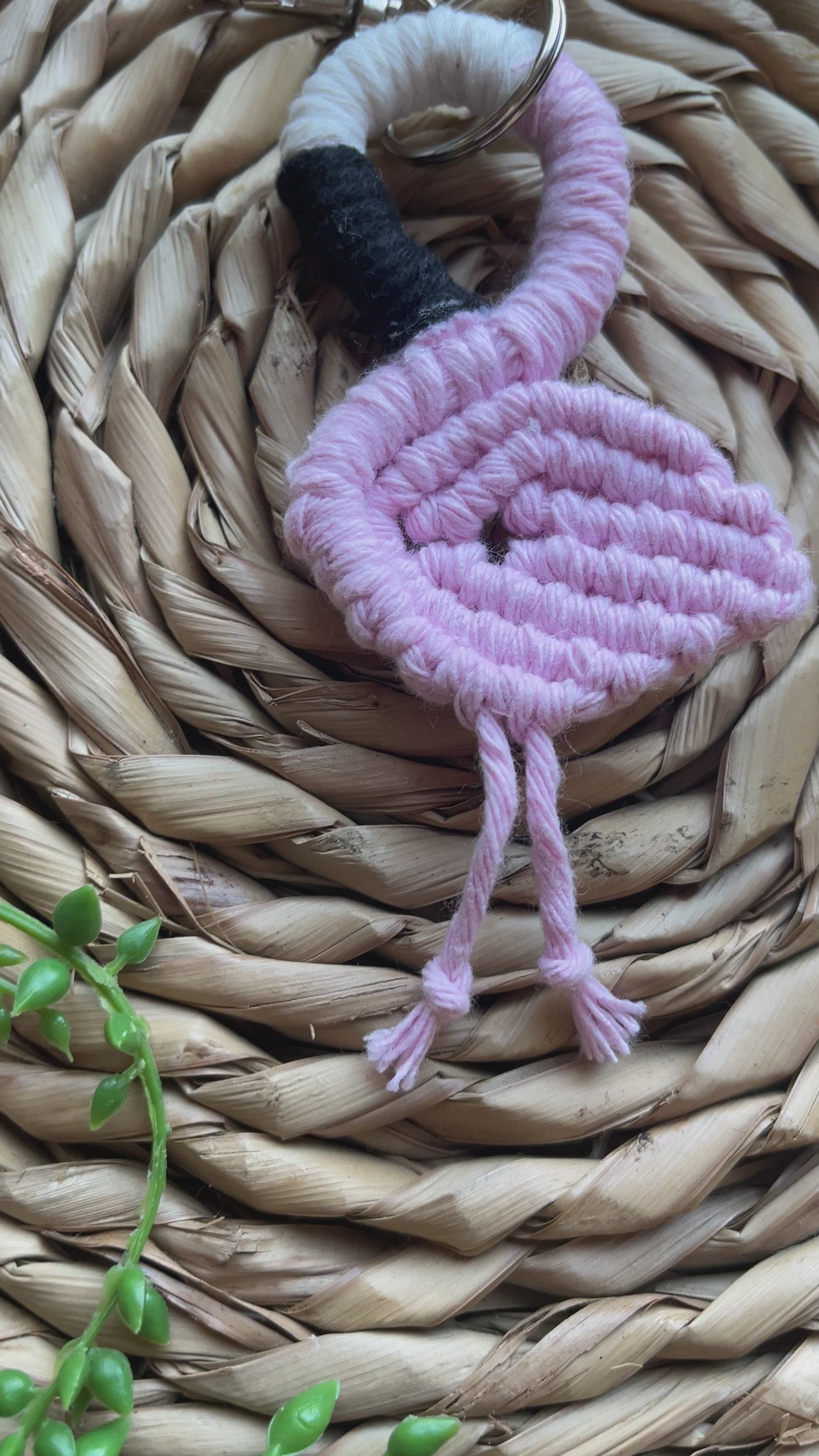 Video showing a handmade macrame keychain in the shape of a flamingo, made using light pink recycled cotton, on a metal split ring with lobster clasp. The keyring is displayed on a round wicker surface next to some green plant vines.