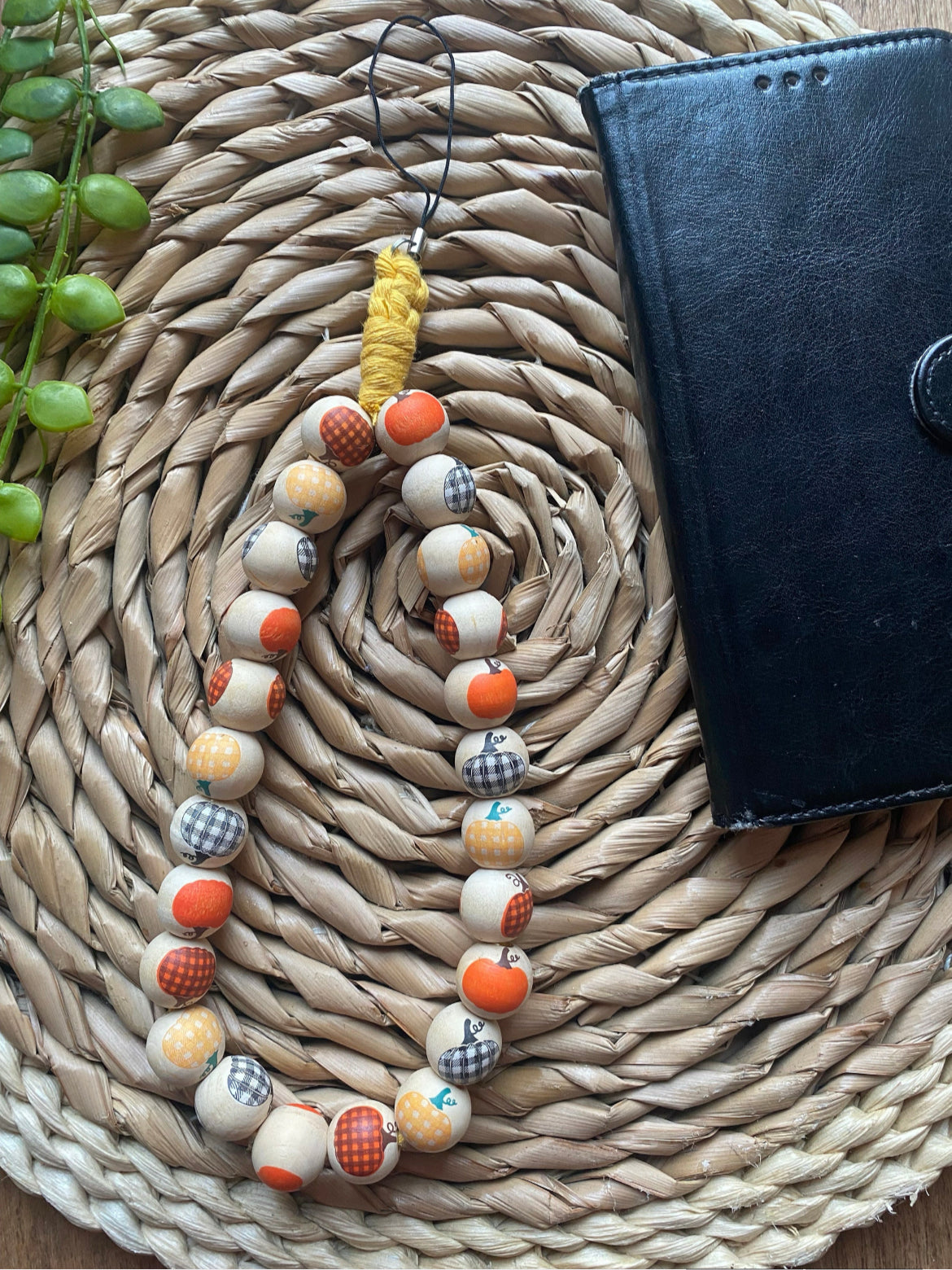 A handmade wristlet phone charm, made with pumpkin patterned wooden beads and a universal phone attachment, on a wicker woven placemat next to a black wallet style phone case.