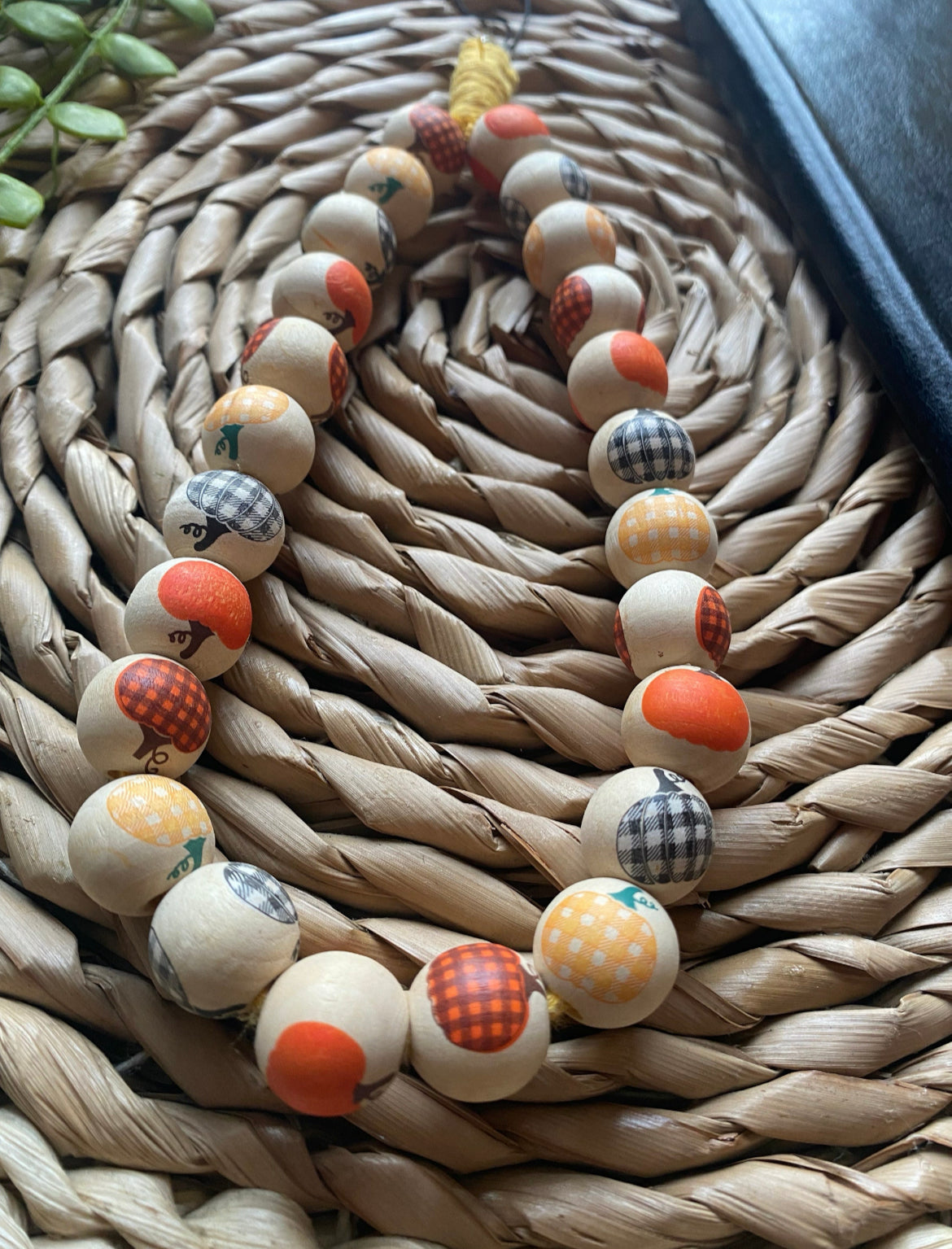 Close up of wooden pumpkin pattern beads on a handmade wristlet phone charm, on a wicker woven placemat next to a black wallet style phone case.