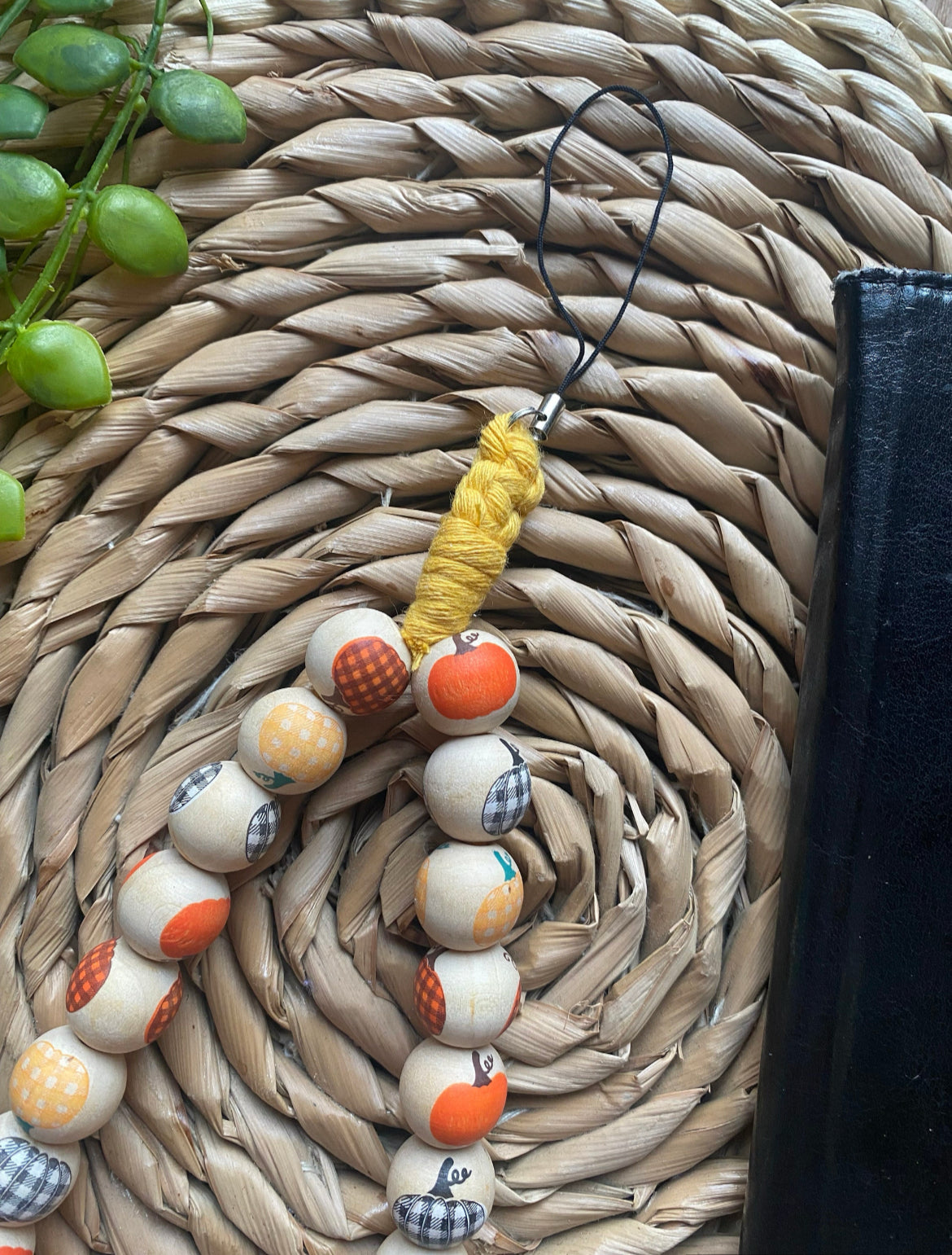 A handmade wristlet phone charm, made with pumpkin patterned wooden beads and a universal phone attachment, on a wicker woven placemat next to a black wallet style phone case.