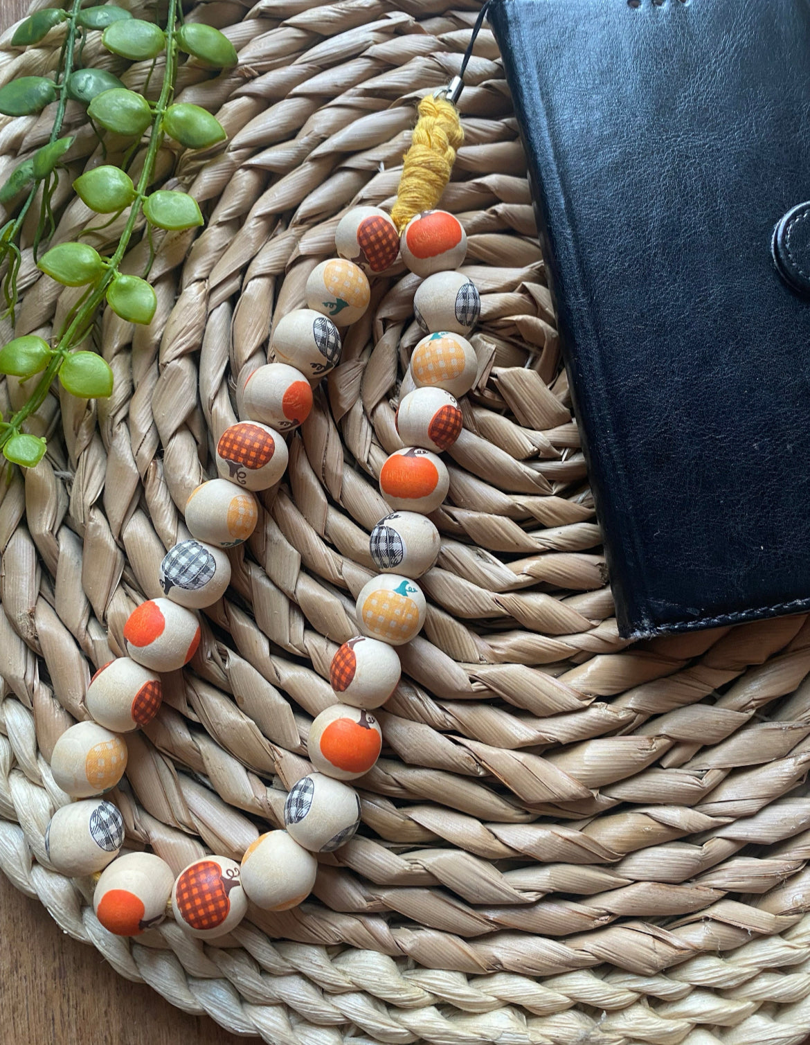A handmade wristlet phone charm, made with pumpkin patterned wooden beads and a universal phone attachment, on a wicker woven placemat attached to a black wallet style phone case.