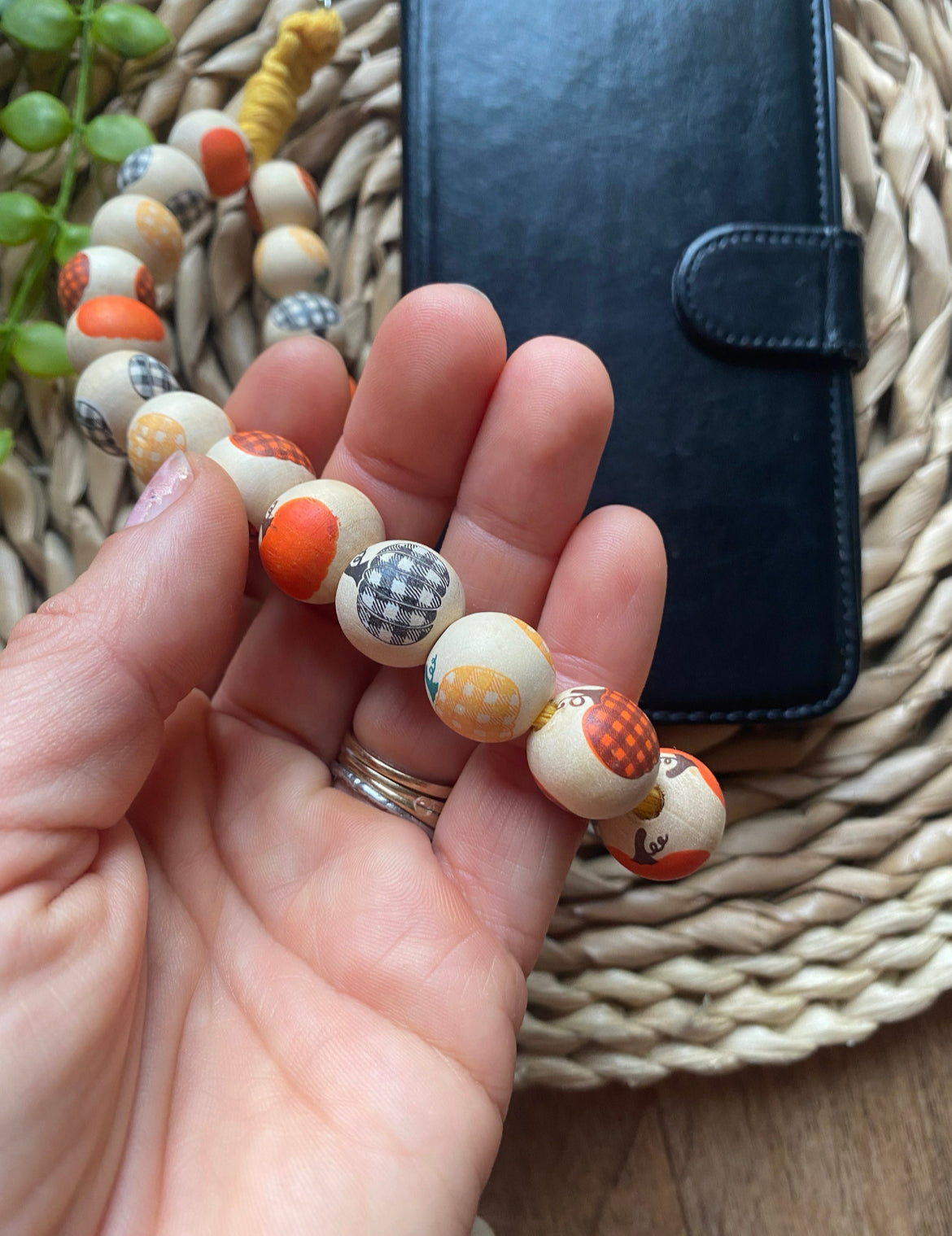 A hand holding a handmade wristlet phone charm, made with pumpkin patterned wooden beads on a wicker woven placemat next to a black wallet style phone case.