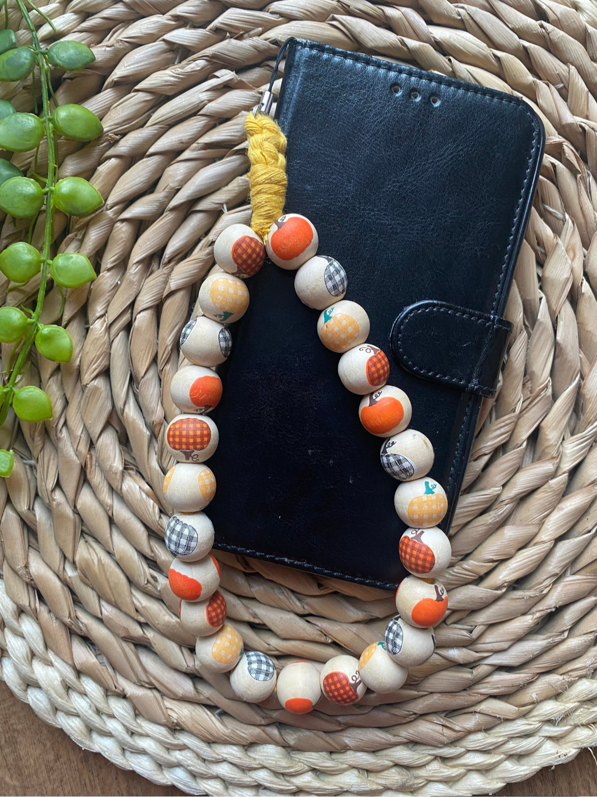 A handmade wristlet phone charm, made with pumpkin patterned wooden beads and a universal phone attachment, laid across a black wallet style phone case.