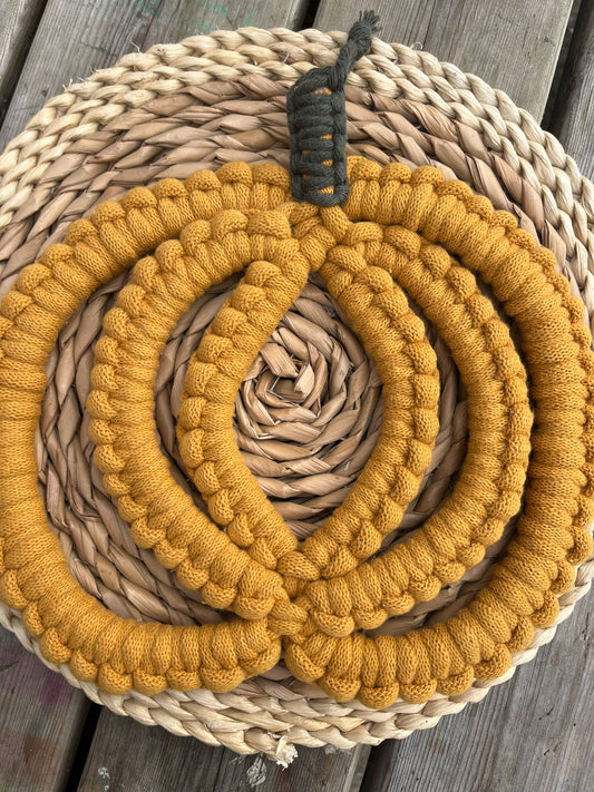 A chunky pumpkin shaped macrame wall hanging made with mustard yellow yarn, displayed on a round wicker mat.