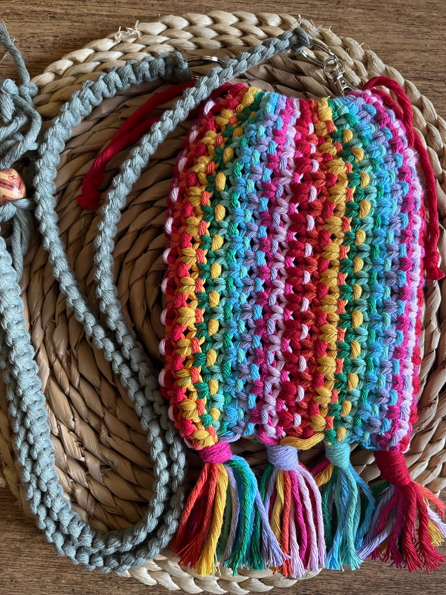 A small macrame bag made with rainbow coloured yarn and a long green strap, on a round wicker mat.