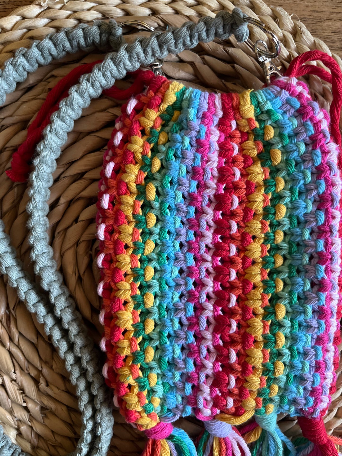 A macrame crossbody bag, made with rainbow yarn and a green strap, on a wicker mat.