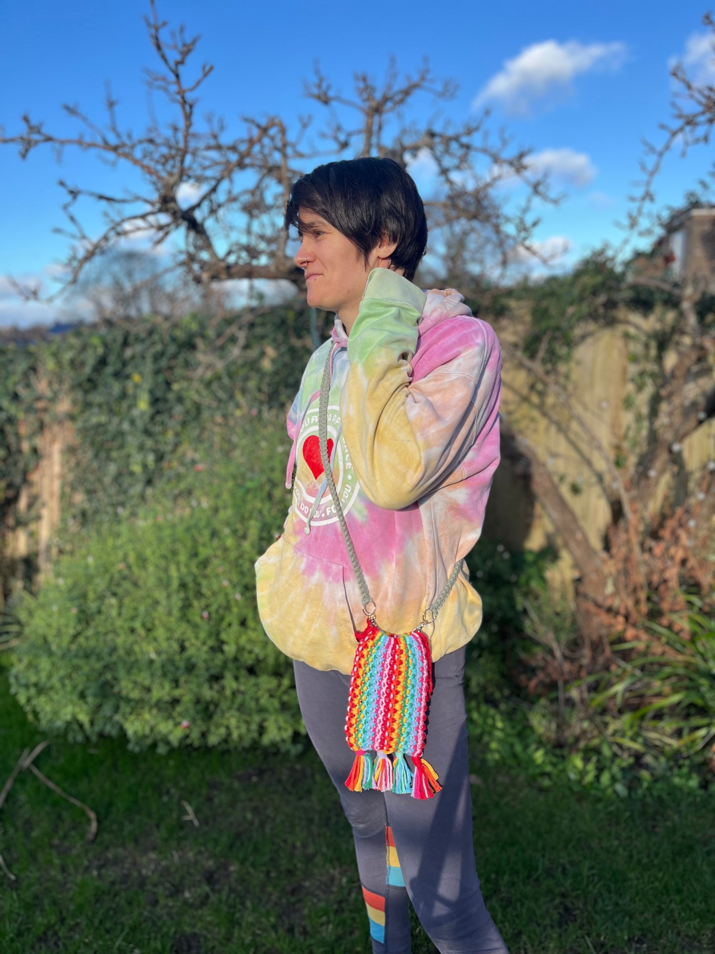 A woman stands outside wearing a rainbow hoodie, facing sideways on to the camera. She is wearing a crossbody bag, which is made with rainbow coloured yarn and a long green strap.