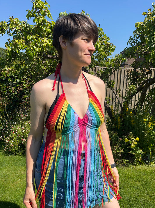 A person stands outdoors on a sunny day, wearing a colorful Rainbow coverup top by Macra-Made-With-Love. They have short dark hair and are looking off to the side. The background features green foliage and a wooden fence, with a bright blue sky overhead.