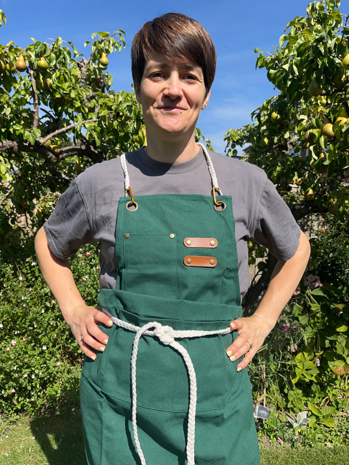 A person with short brown hair stands outside with their hands on their hips, they are wearing a green apron with a white braided rope tie.