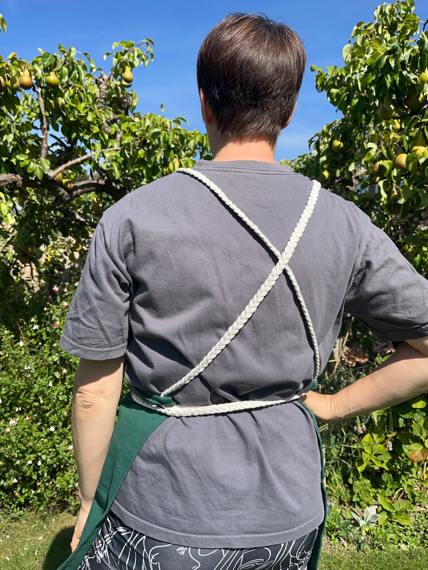 A person with short brown hair stands outside with their back to the camera, they are wearing a grey t shirt & the cross back braided straps of their apron are visible.