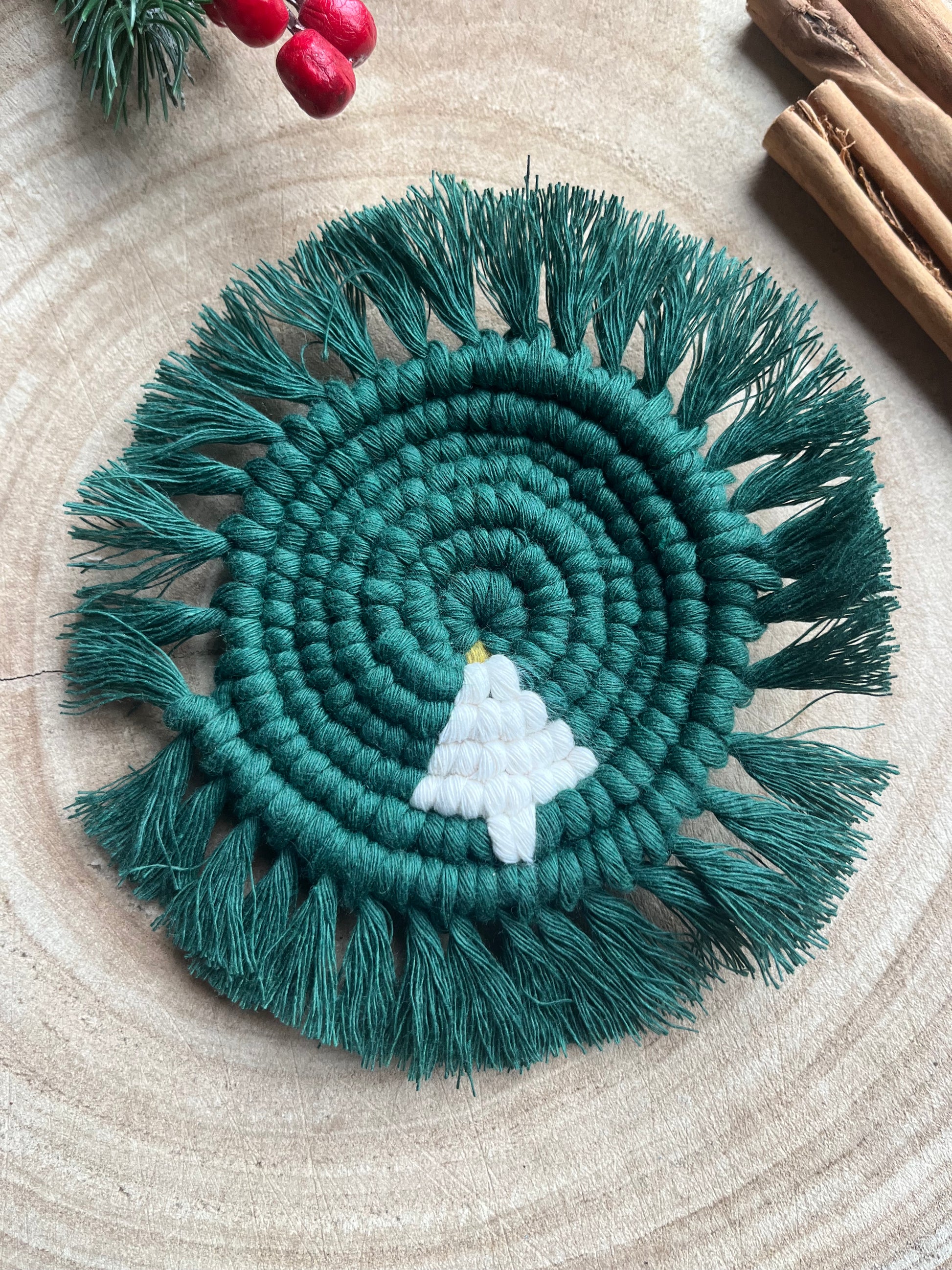 A handmade macrame round coaster with a Christmas Tree detail in the centre, and fringed edge, displayed on a wooden surface.