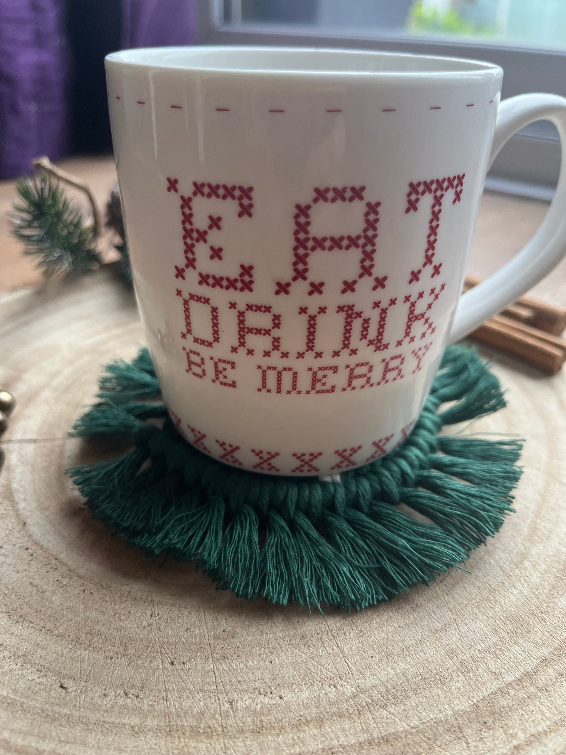 A white mug with red text sits on top of a round macramé coaster made with green yarn, with a fringed edge.