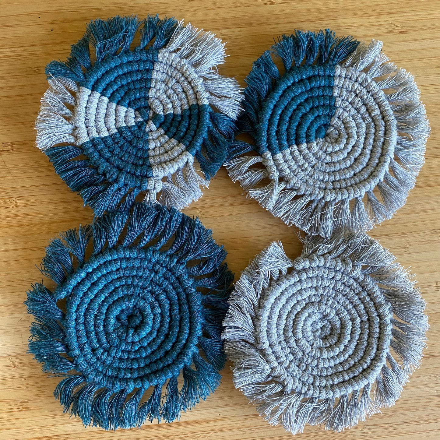 A set of 4 round macrame coasters made with blue and grey yarns, with a soft fringed edge, are placed on a wooden table.