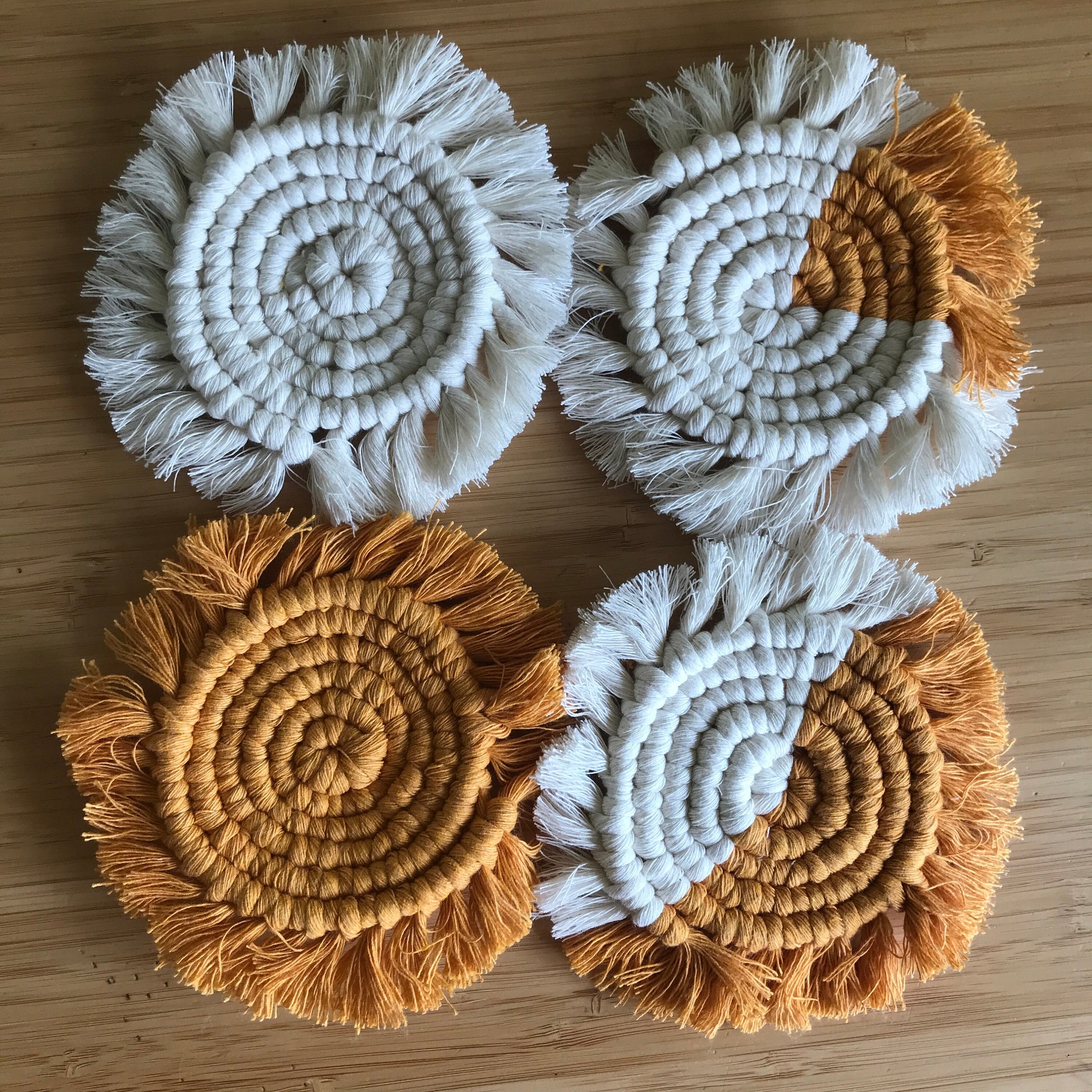 A set of 4 round macrame coasters made with mustard yellow and natural yarns, with a soft fringed edge, are placed on a wooden table.